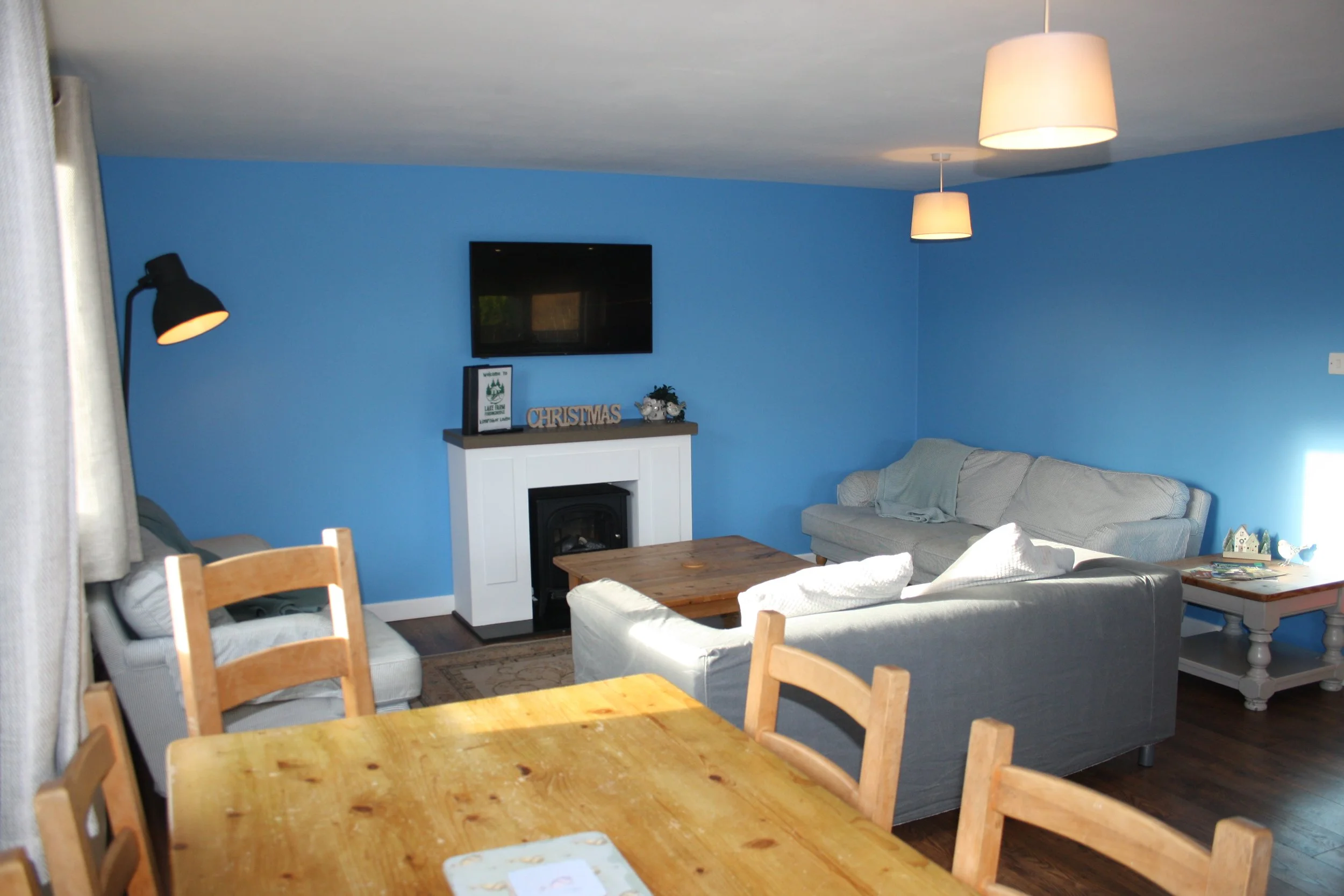Living room with blue walls, a wall-mounted TV, a white fireplace with decorations including a framed picture and a 'Christmas' sign, a gray sofa, a wooden coffee table, a side table with a house-shaped decor, a curtain on the left, and ceiling lights.