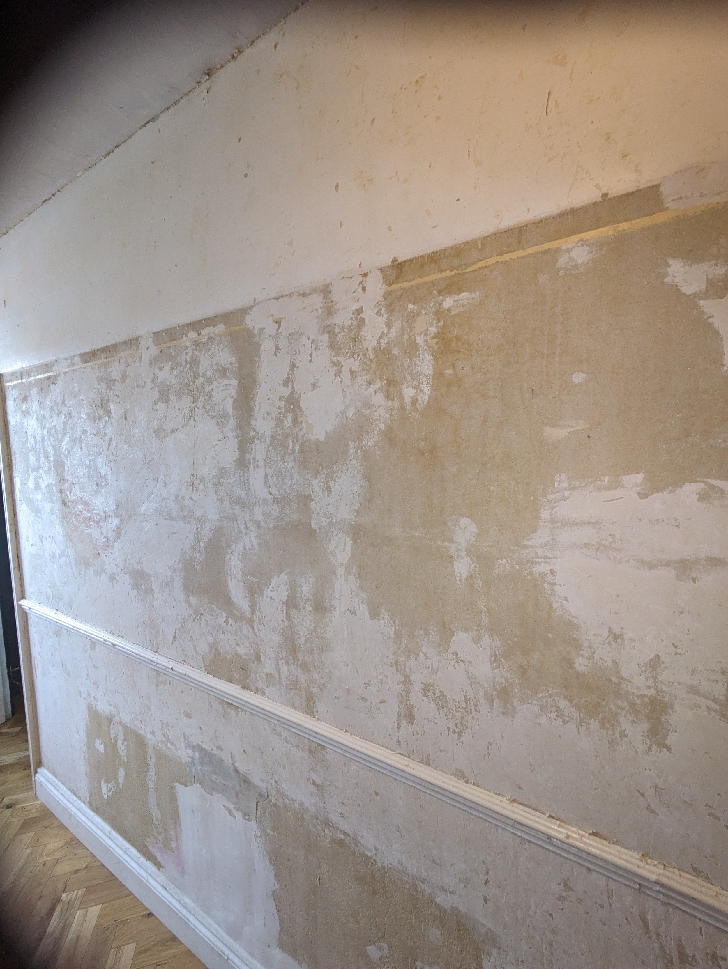 A wall with a partially removed textured beige wallpaper and a decorative white molding at the bottom, with flooring visible at the bottom of the image.
