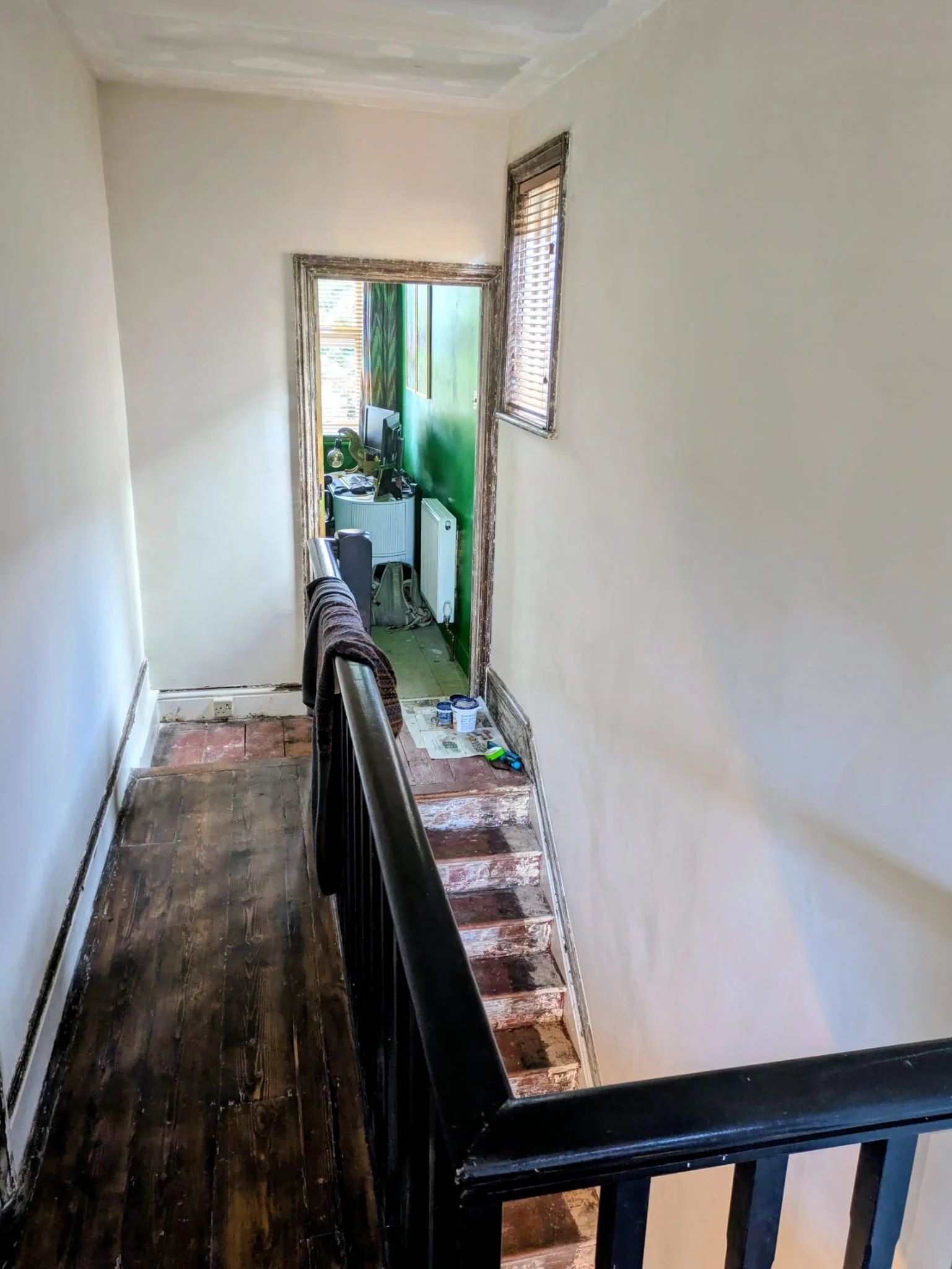 Interior view of a hallway with dark wooden flooring, white walls, and a staircase with a black railing leading to another room, which has a green wall and windows with blinds.