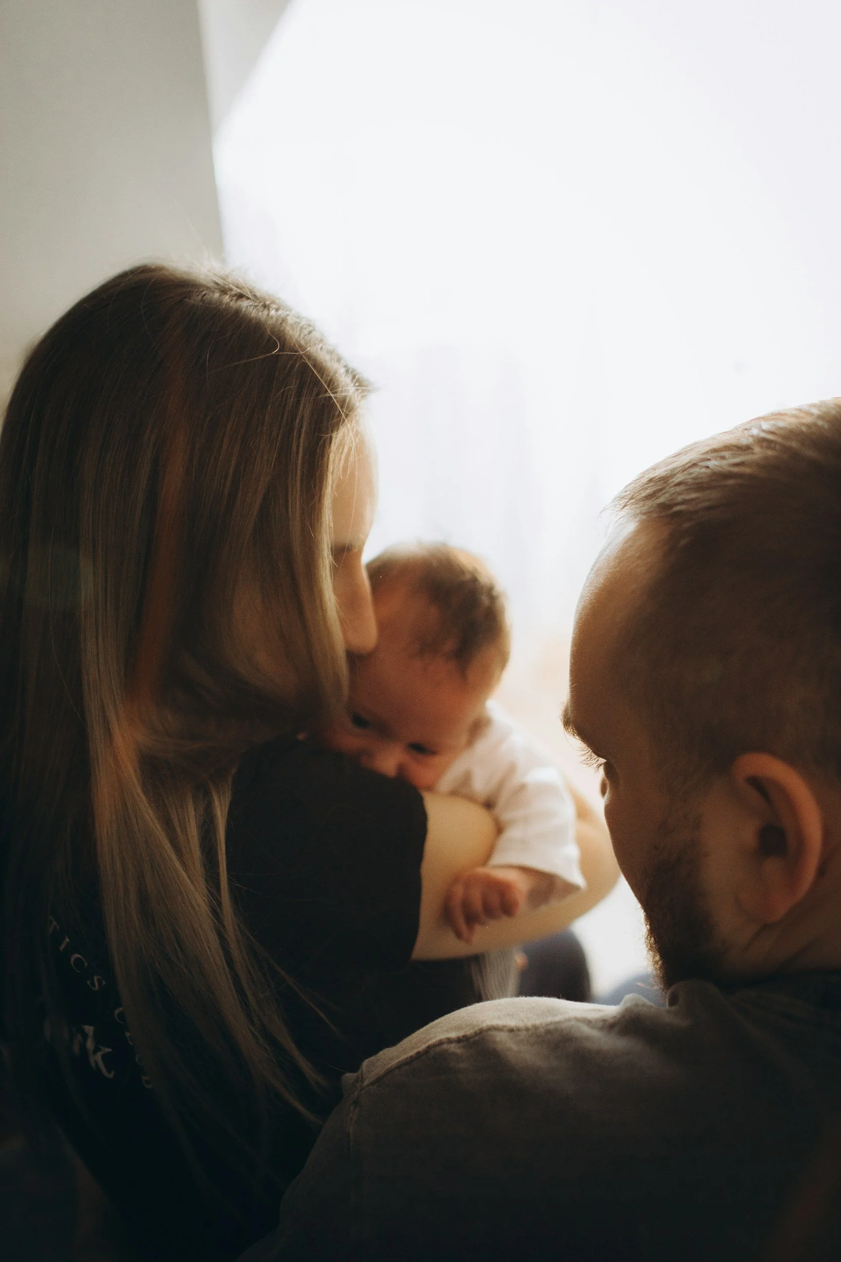 A woman with long, light brown hair holds a baby while sharing a close moment with a man with a shaved head and beard. The scene is intimate, with the baby gazing at the man and the woman smiling softly.