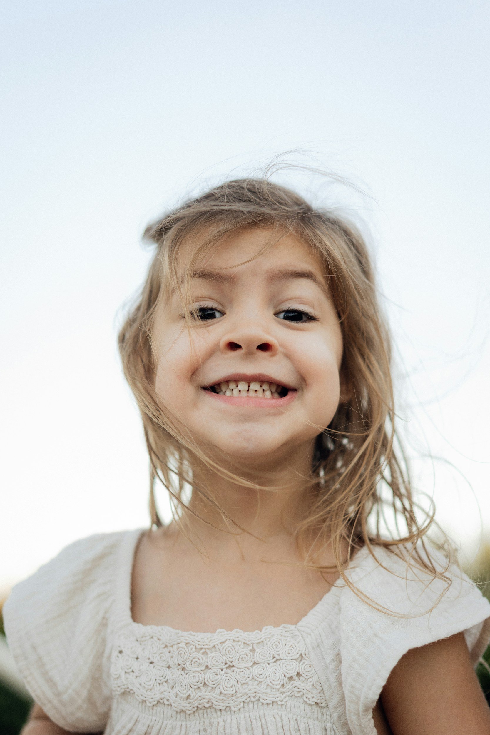 Close-up of a young girl smiling outdoors, wearing a white dress with detailed embroidery.