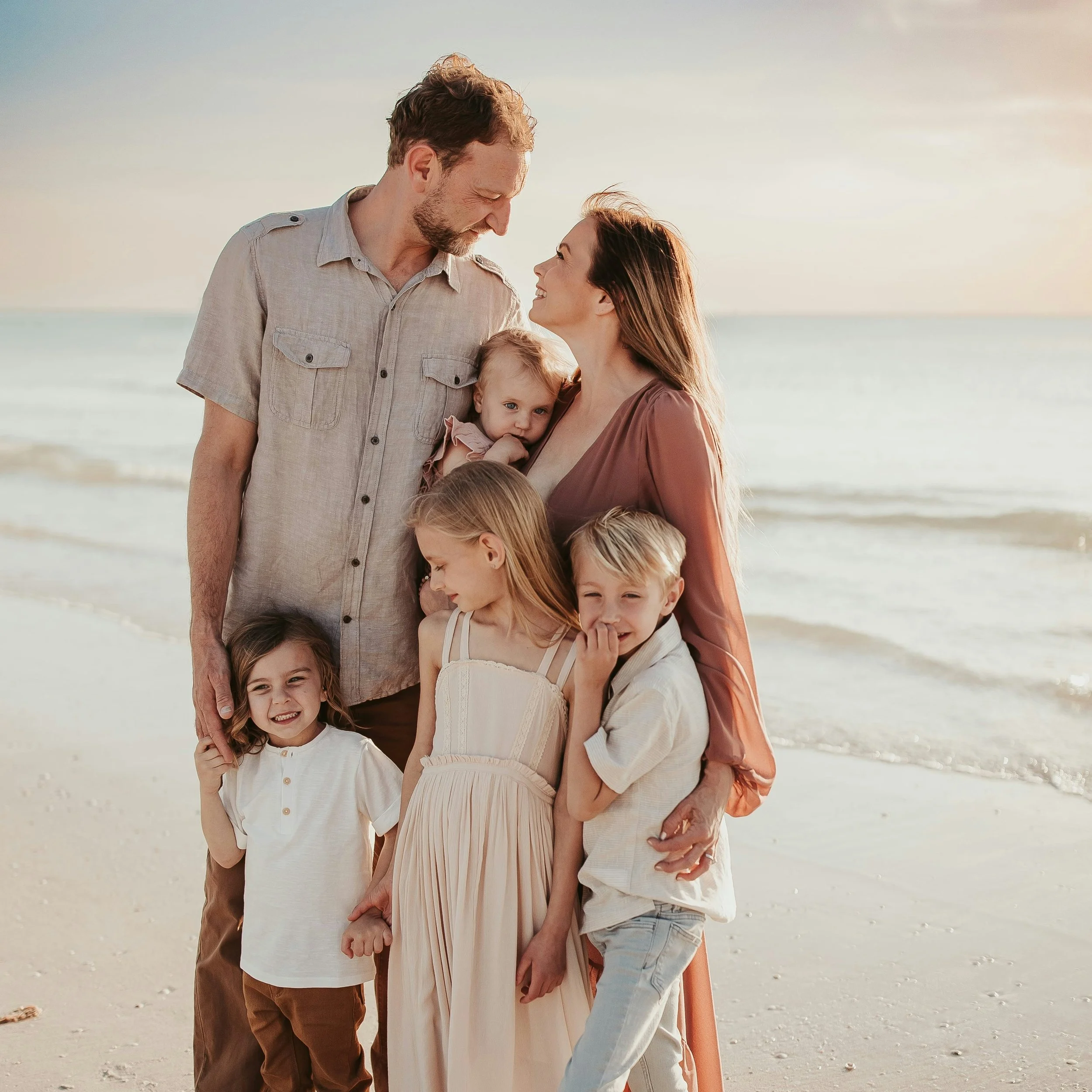 A happy family of six, including two adults and four children, stands close together on the beach during sunset, smiling and looking at each other.