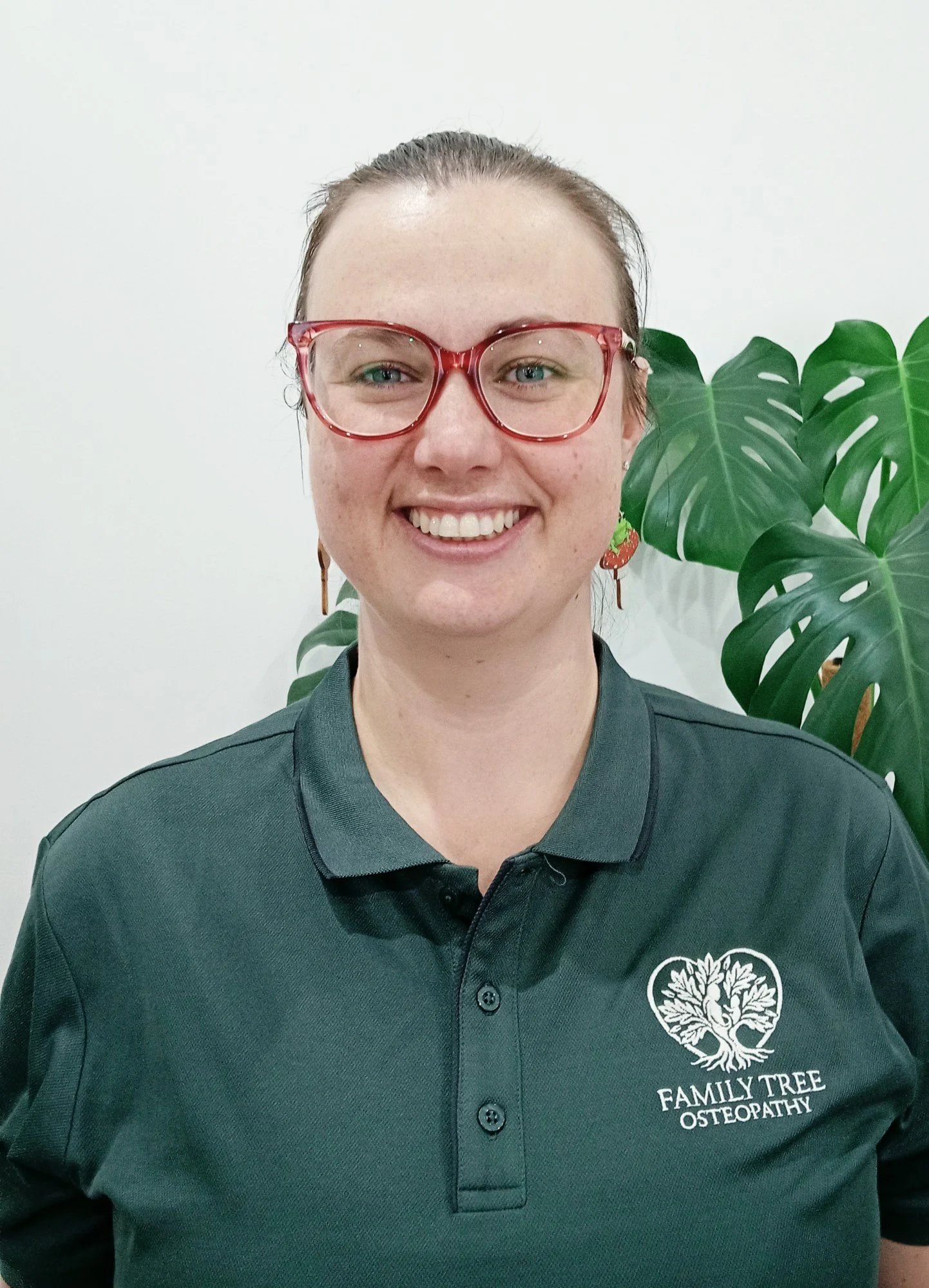 A woman with glasses and earrings smiling in front of a large green plant, wearing a dark green polo shirt with a Family Tree Osteopathy logo.