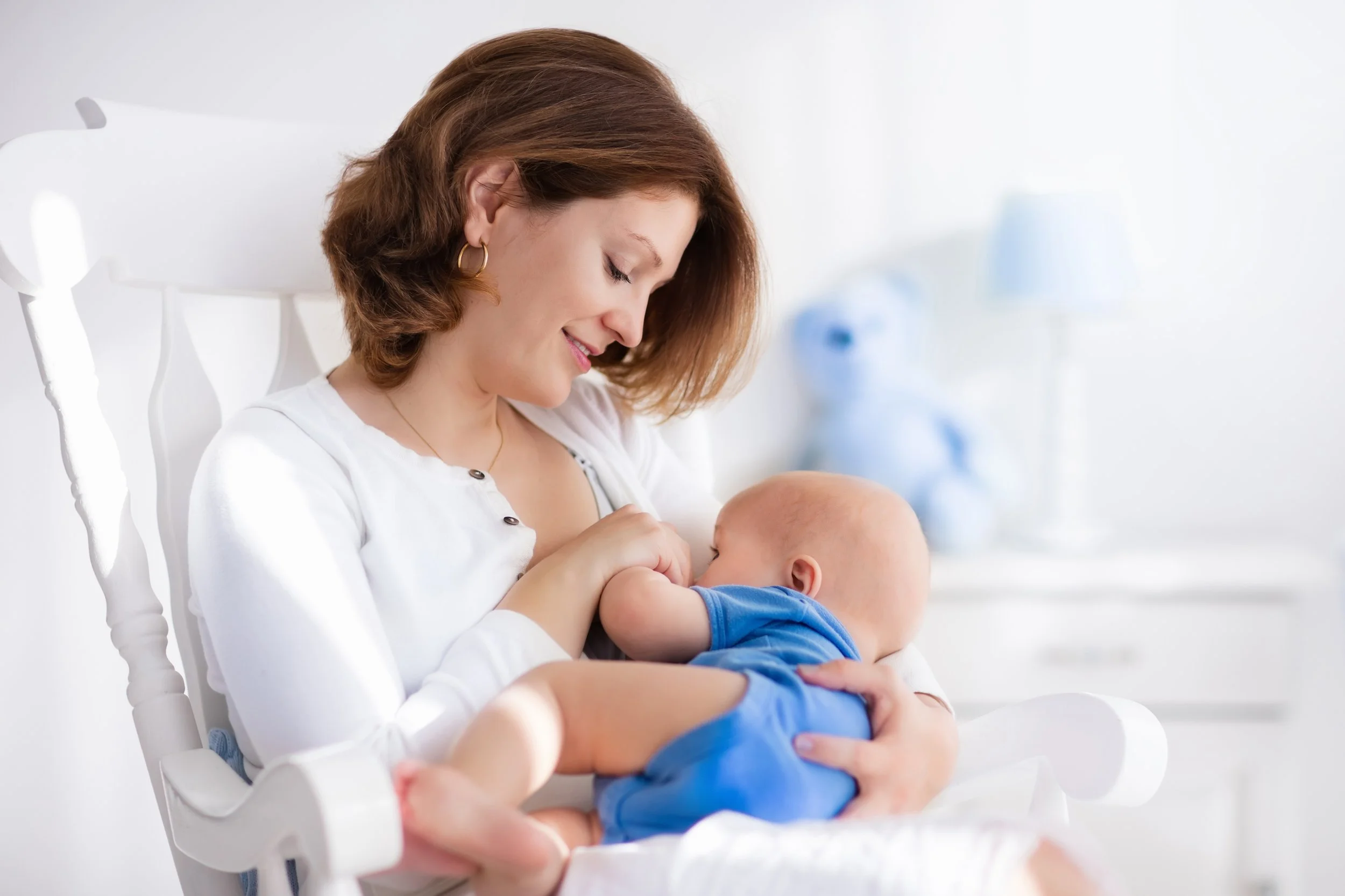 A woman with short brown hair wearing earrings and a white shirt is smiling and gently holding a baby in her arms, looking at the child affectionately.