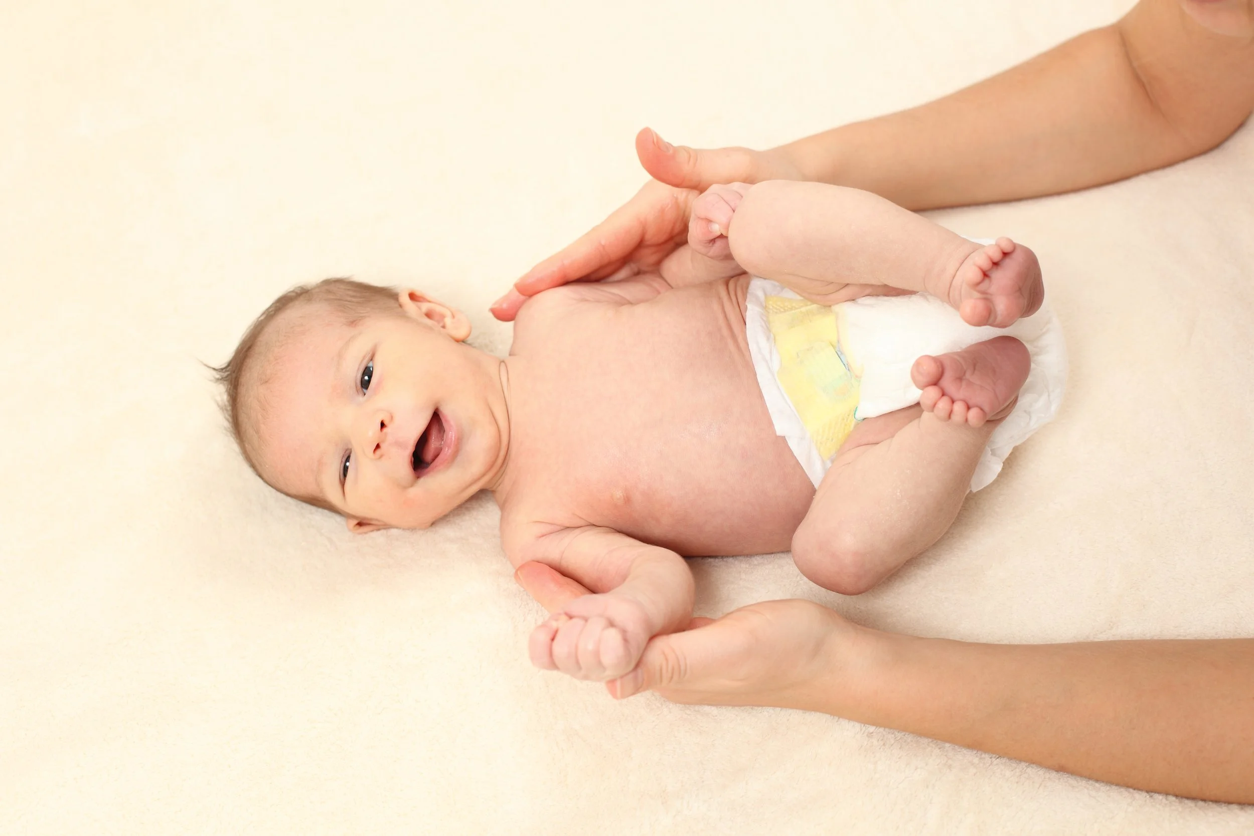 A smiling baby lying on a soft surface with an adult holding the baby's legs and feet, the baby's face is expressive and cheerful.