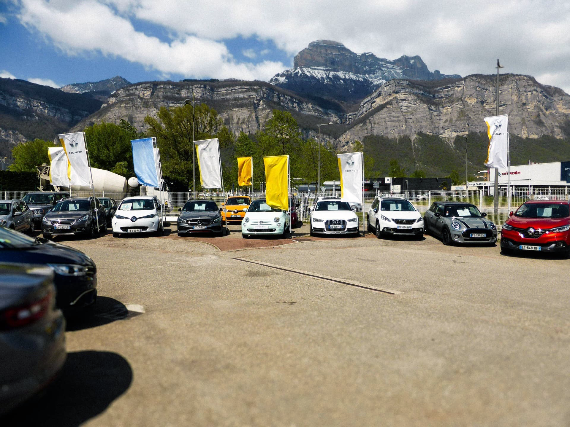 Un parking avec plusieurs voitures et des drapeaux blancs et jaunes, avec en arrière-plan une grande montagne rocheuse au sommet enneigé et des arbres verts.