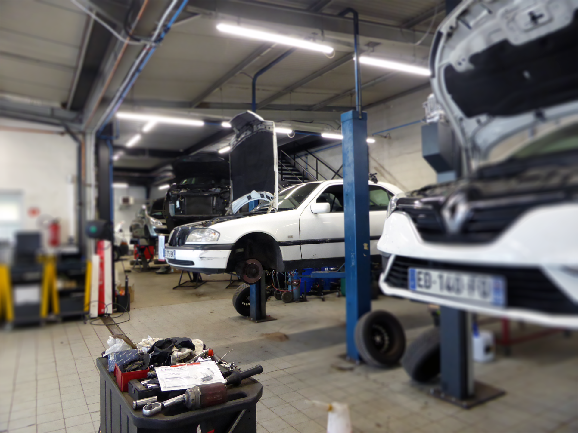 Garage de réparation automobile avec plusieurs voitures en cours de réparation, notamment une voiture blanche sur un lift, un moteur ouvert, et des outils dispersés au sol.