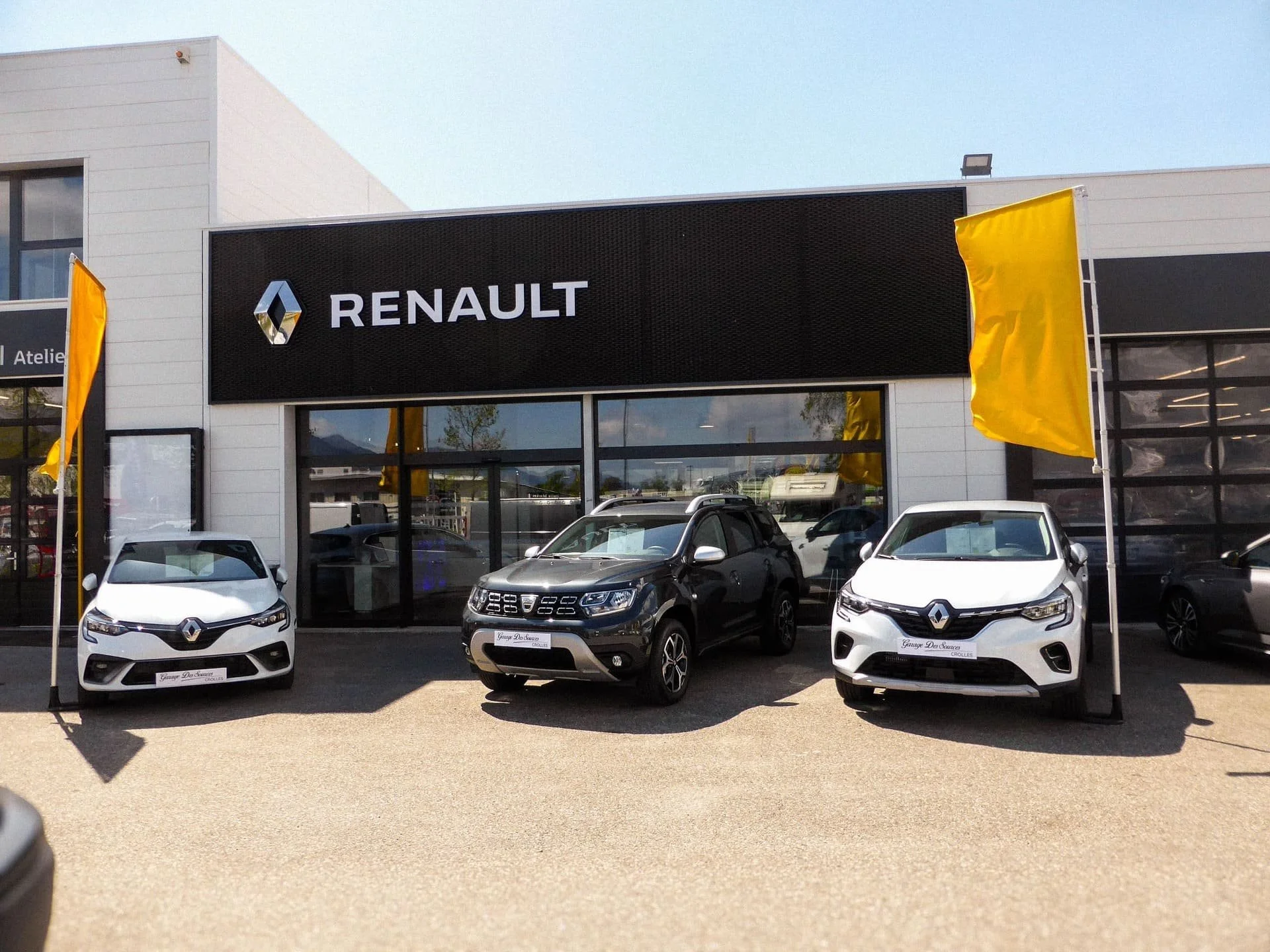 Une concession automobile Renault avec plusieurs voitures garées à l'extérieur et deux drapeaux jaunes.