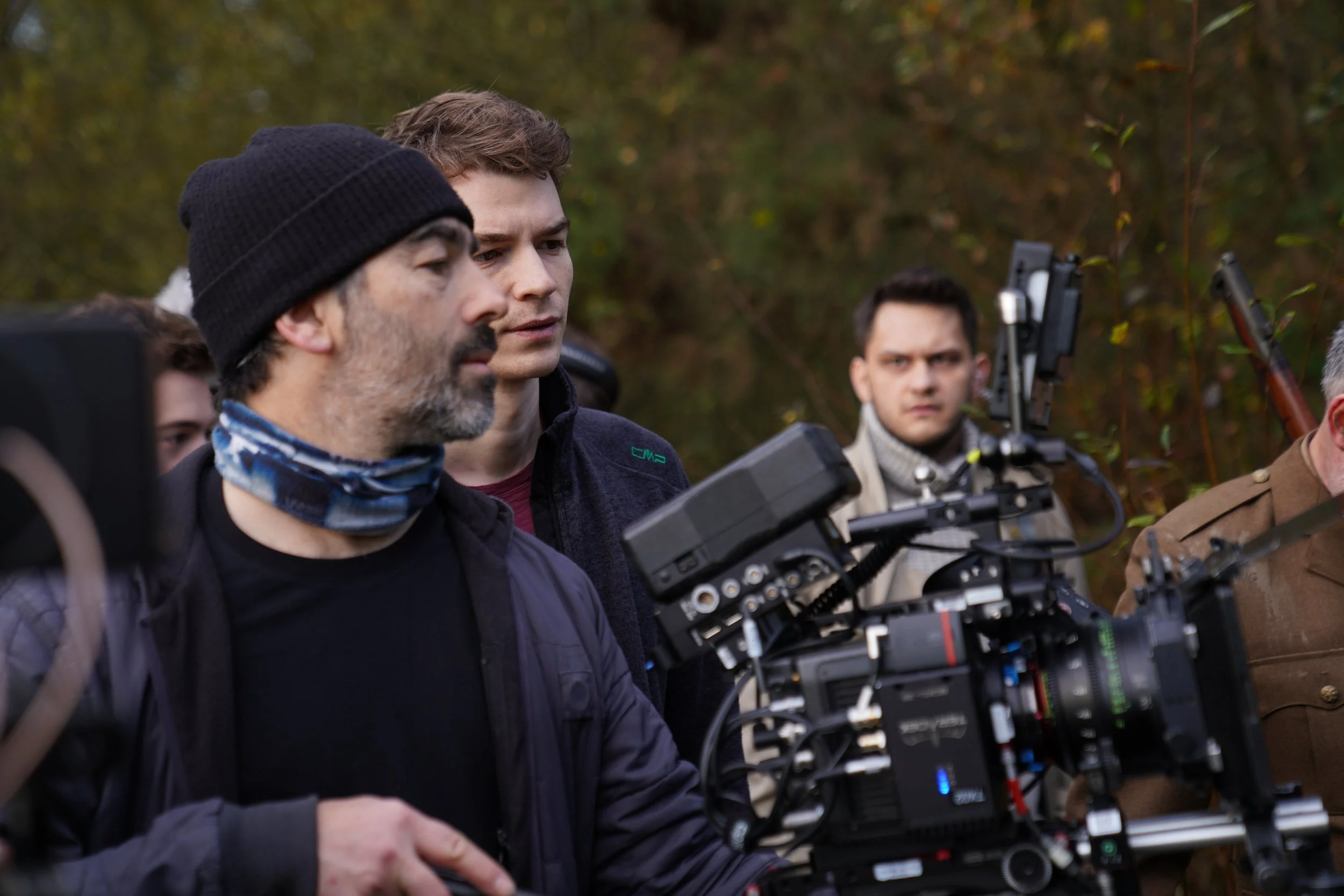 Multiple people, including a man wearing a black beanie and another man with curly hair, are gathered outdoors around a professional film camera, with trees in the background.