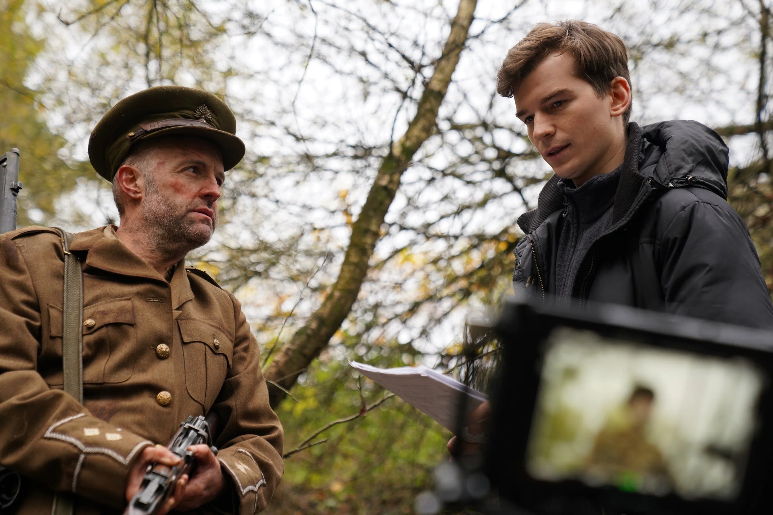A man dressed as a soldier from the early 20th century, wearing a brown uniform and hat, holds a pistol, talking to a younger man in a black jacket outdoors with autumn trees.