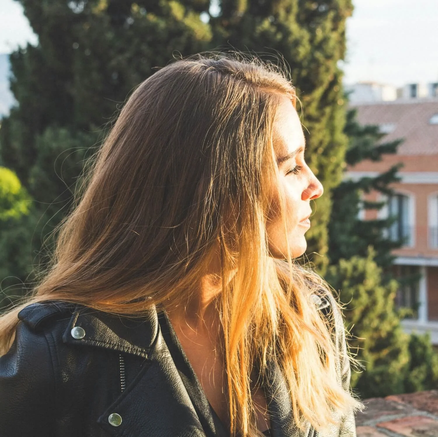 Seitenansicht einer jungen Frau mit langen, hellbraunen Haaren, die die Augen geschlossen hat und Sonnenlicht auf ihrem Gesicht spürt, im Freien mit Bäumen und Gebäuden im Hintergrund.