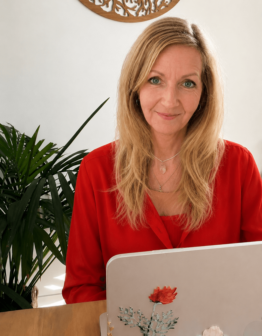 Madeleine Gutmann, in roten Oberteil, sitzt vor einem weißen Hintergrund neben einer großen grünen Pflanze, schaut auf einen Laptop