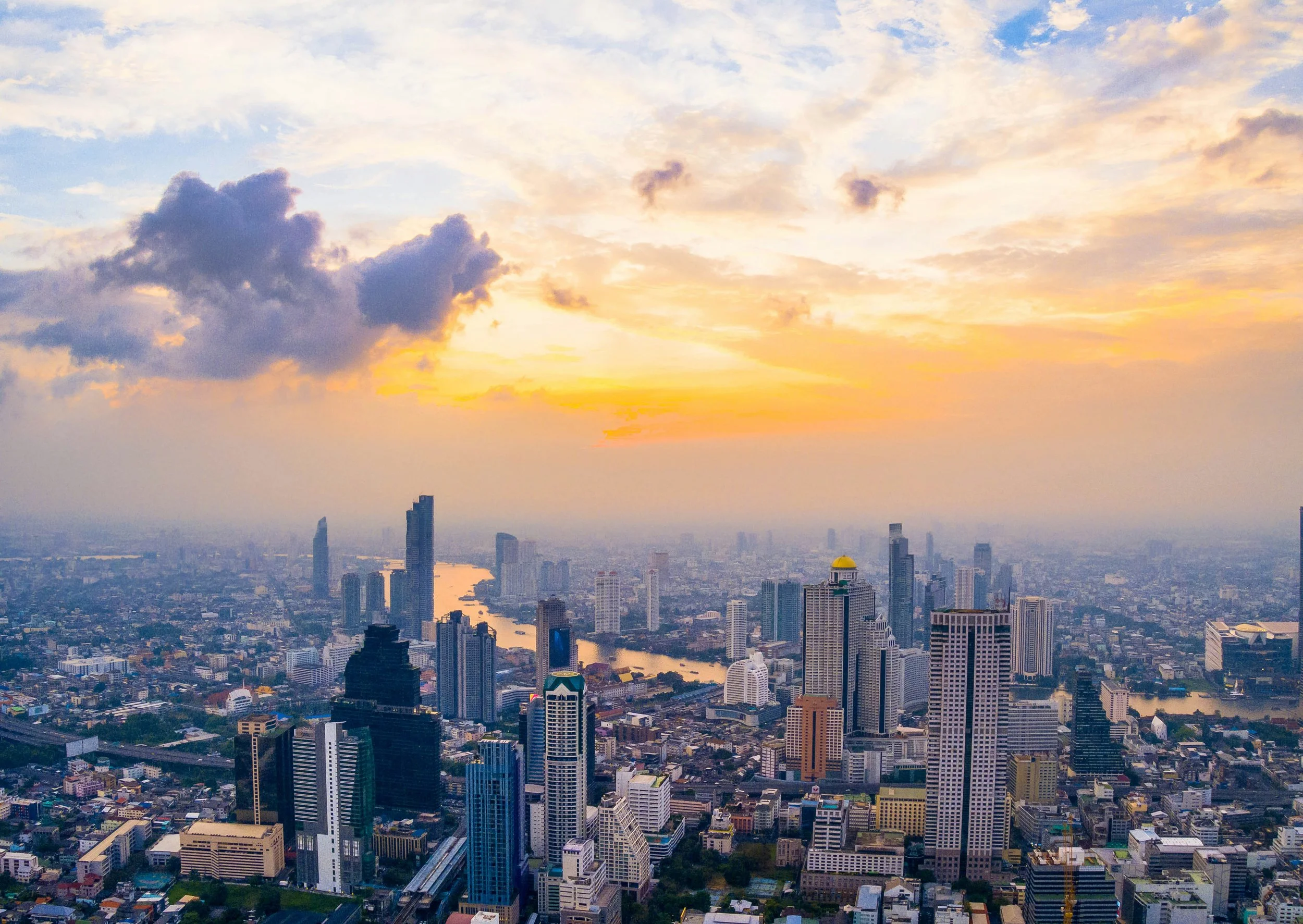A city skyline at sunset with tall skyscrapers, a river running through the city, and a partly cloudy sky with the sun setting in the distance.