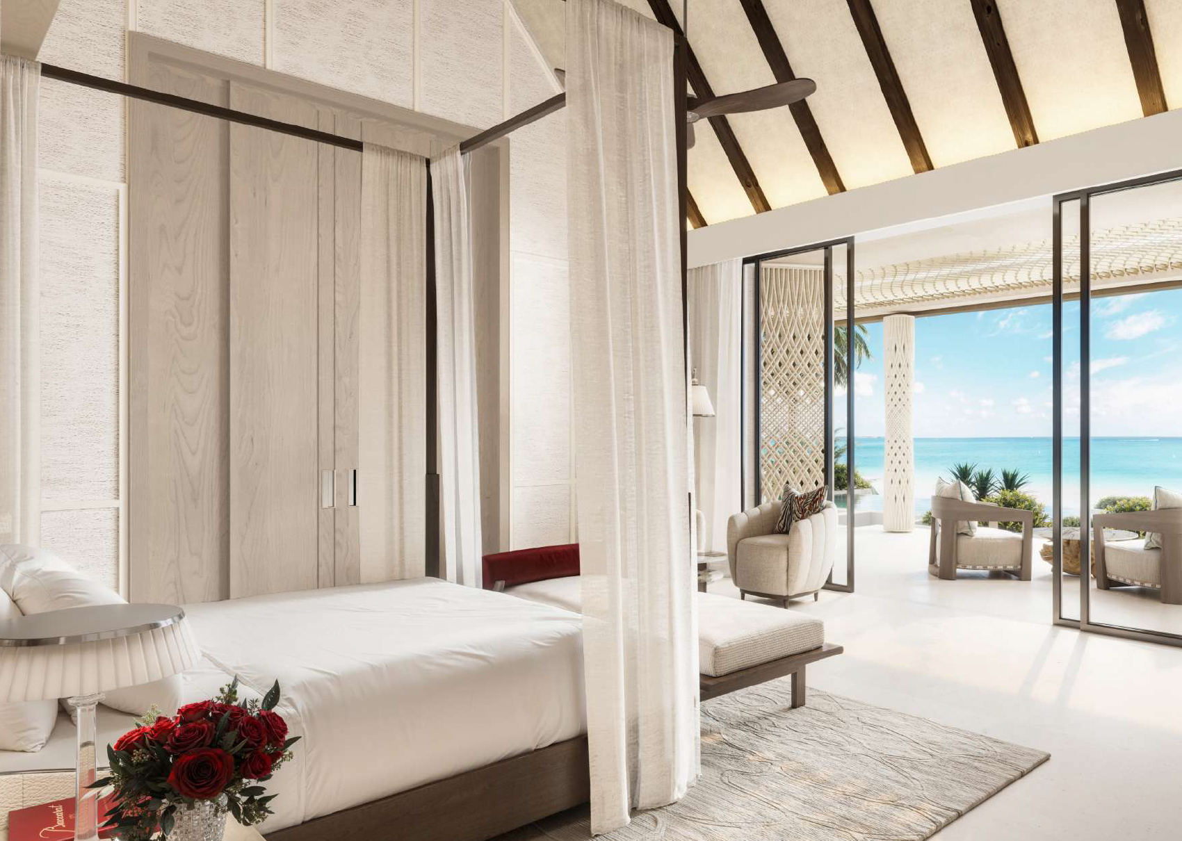 A luxurious beachfront hotel room with a large bed, bedside table with a lamp, and a bouquet of red roses. The room has light-colored walls, curtains, and wooden accents. Large sliding glass doors open to a balcony with outdoor seating and a view of 