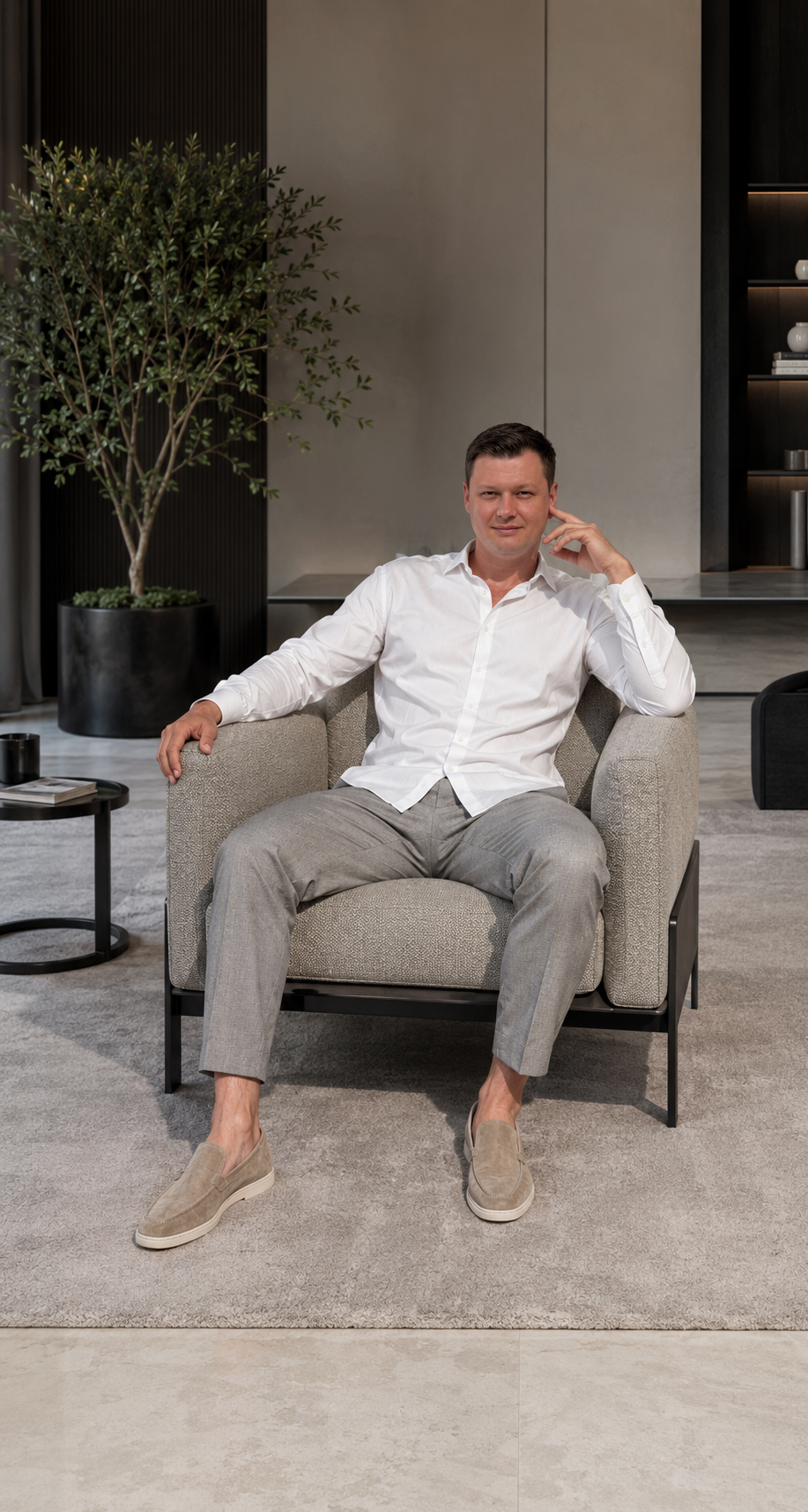 A man sitting in a contemporary living room with a white dress shirt, gray pants, and beige loafers. He is sitting on a light-colored sofa, with a large potted tree and modern decor visible in the background.
