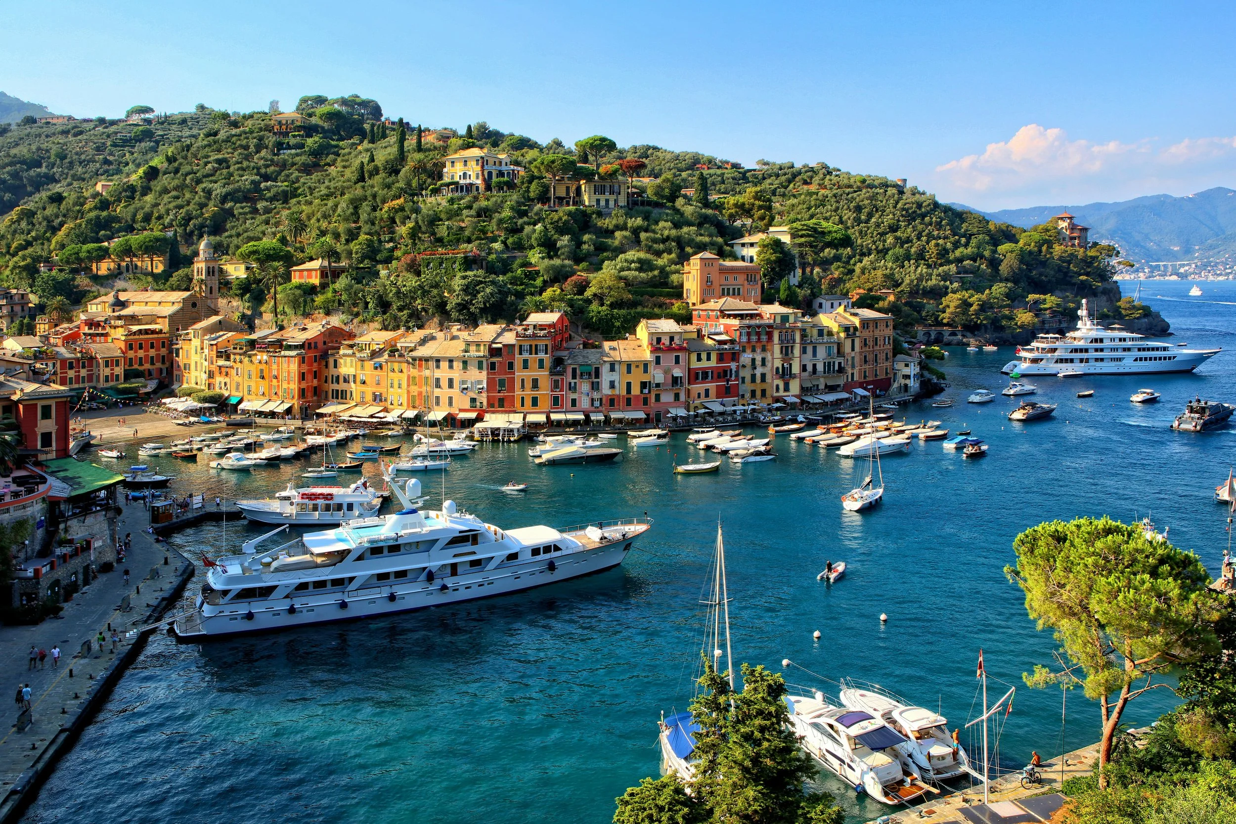 Colorful buildings along a waterfront with boats and yachts in the harbor, lush green hillside with houses and trees, mountains in the background, bright blue sky.