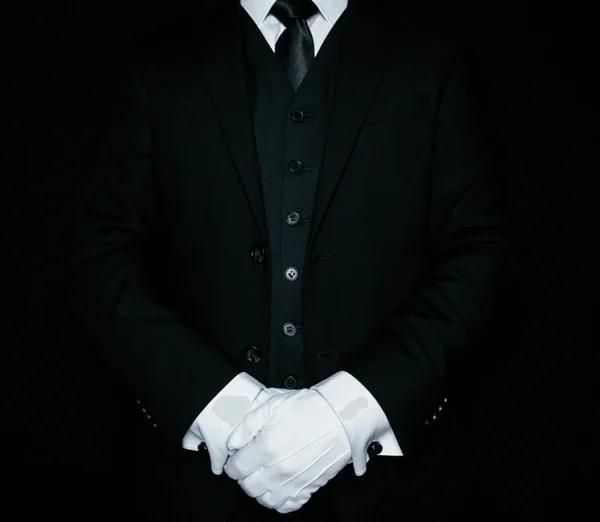 Person dressed in a black suit with a white shirt and white gloves, standing with hands clasped in front against a dark background.