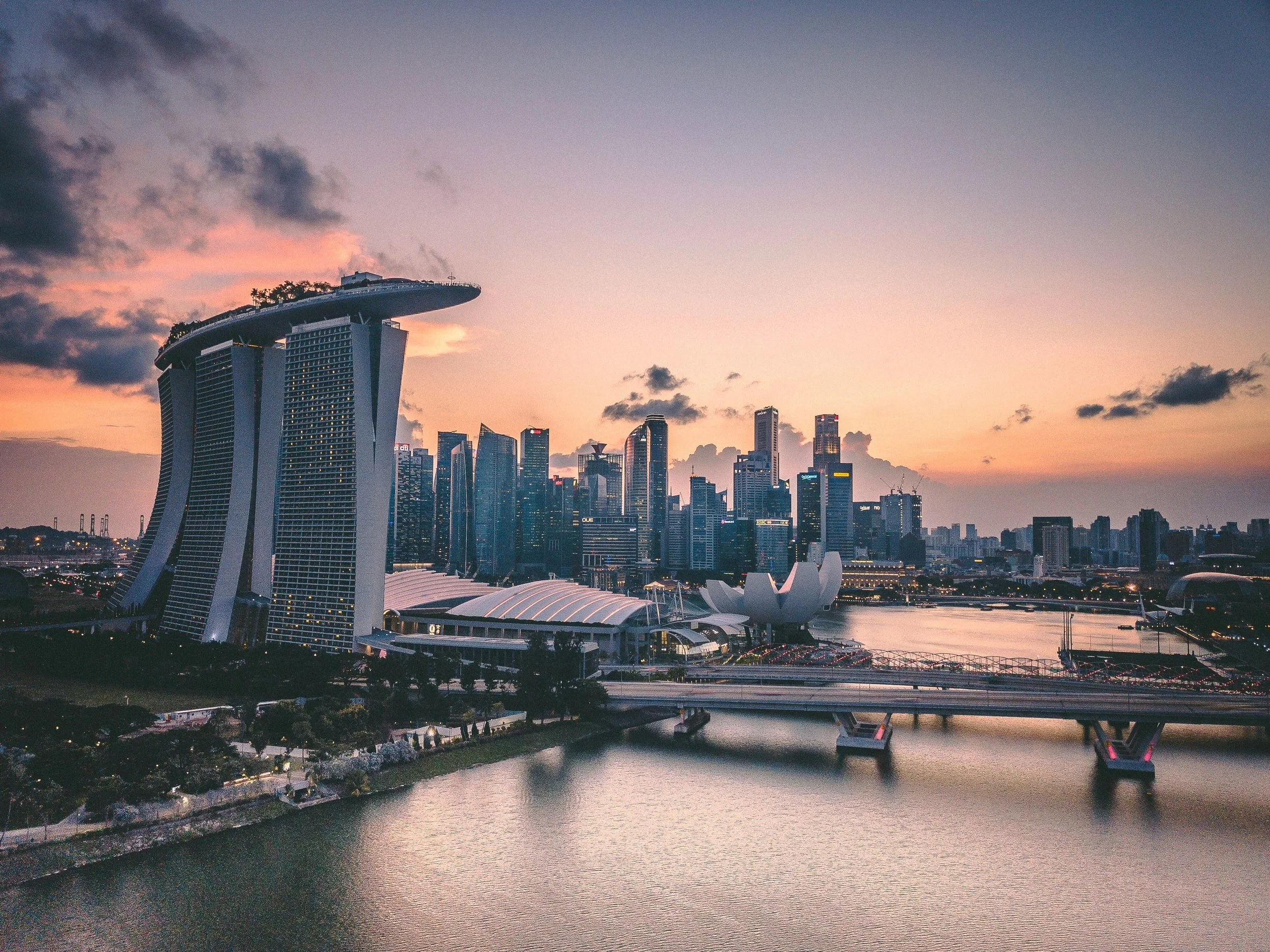 A city skyline at sunset featuring the Marina Bay Sands hotel with three towers and a boat-shaped rooftop. The skyline includes modern skyscrapers, and there is a river in the foreground with a bridge crossing over it.