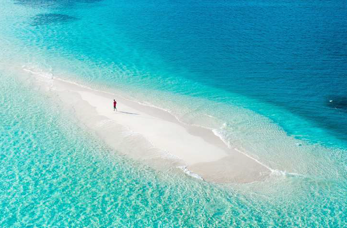 A person standing on a narrow sandbar surrounded by clear blue ocean water.