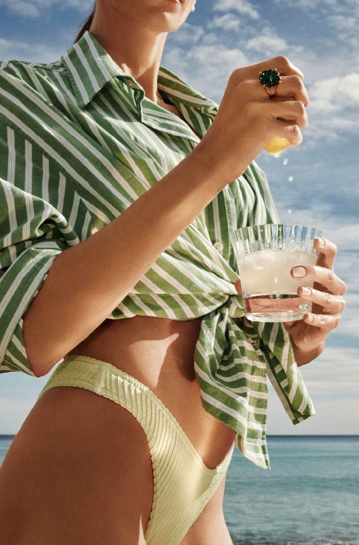 Woman in green and white striped shirt at the beach, holding a glass of a drink and squeezing a lemon.