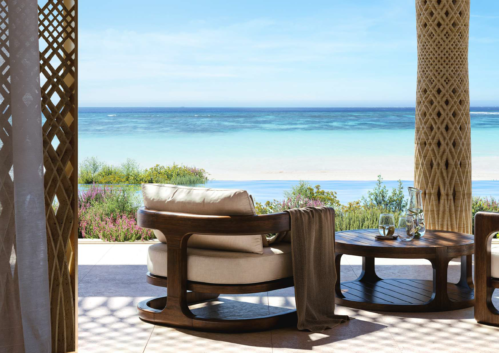 A cozy outdoor seating area with a round wooden table, glass pitcher, glasses, and bench overlooking a beach with clear blue water, sandy shore, and colorful plants and bushes.