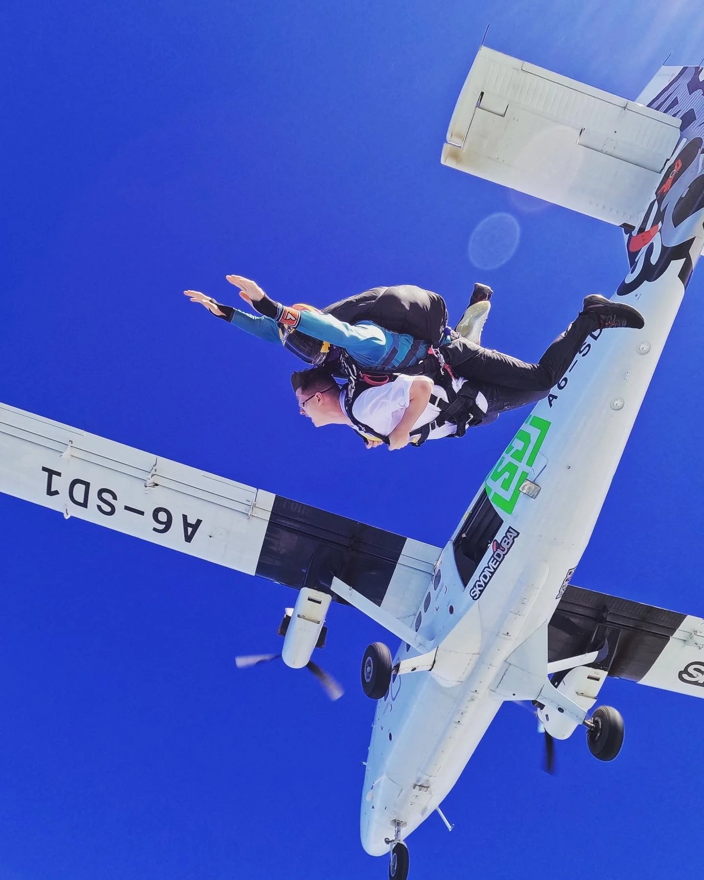A small airplane flying through a bright blue sky with a crew member standing on the wing, wearing a safety harness and looking downward.