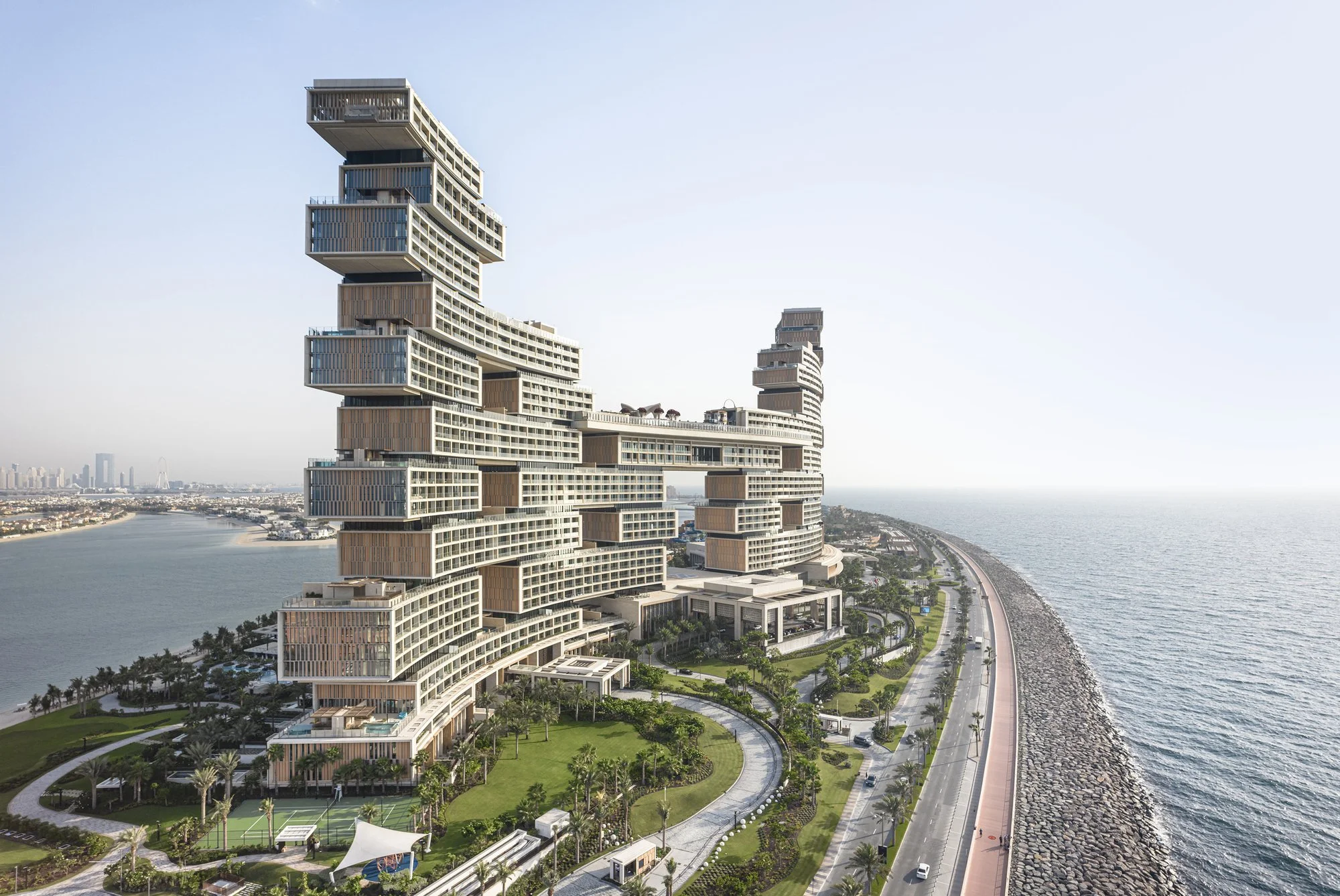 Modern high-rise building with stacked and offset rectangular sections, located along a waterway with a road and rocky shoreline nearby.