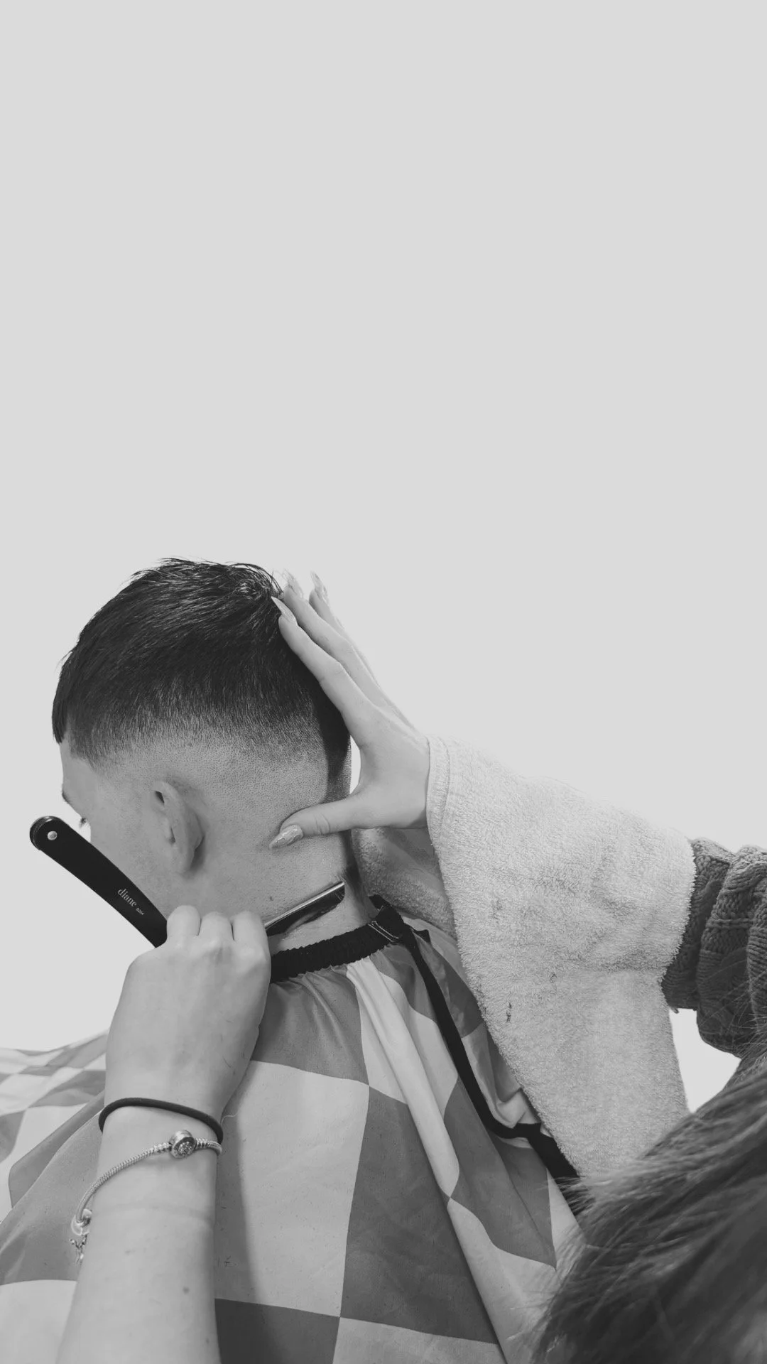 A barber giving a haircut to a young man who is sitting with a cape draped around his shoulders. The barber is using a straight razor to shave or trim the client's neck while holding the client's head steady with her hand.