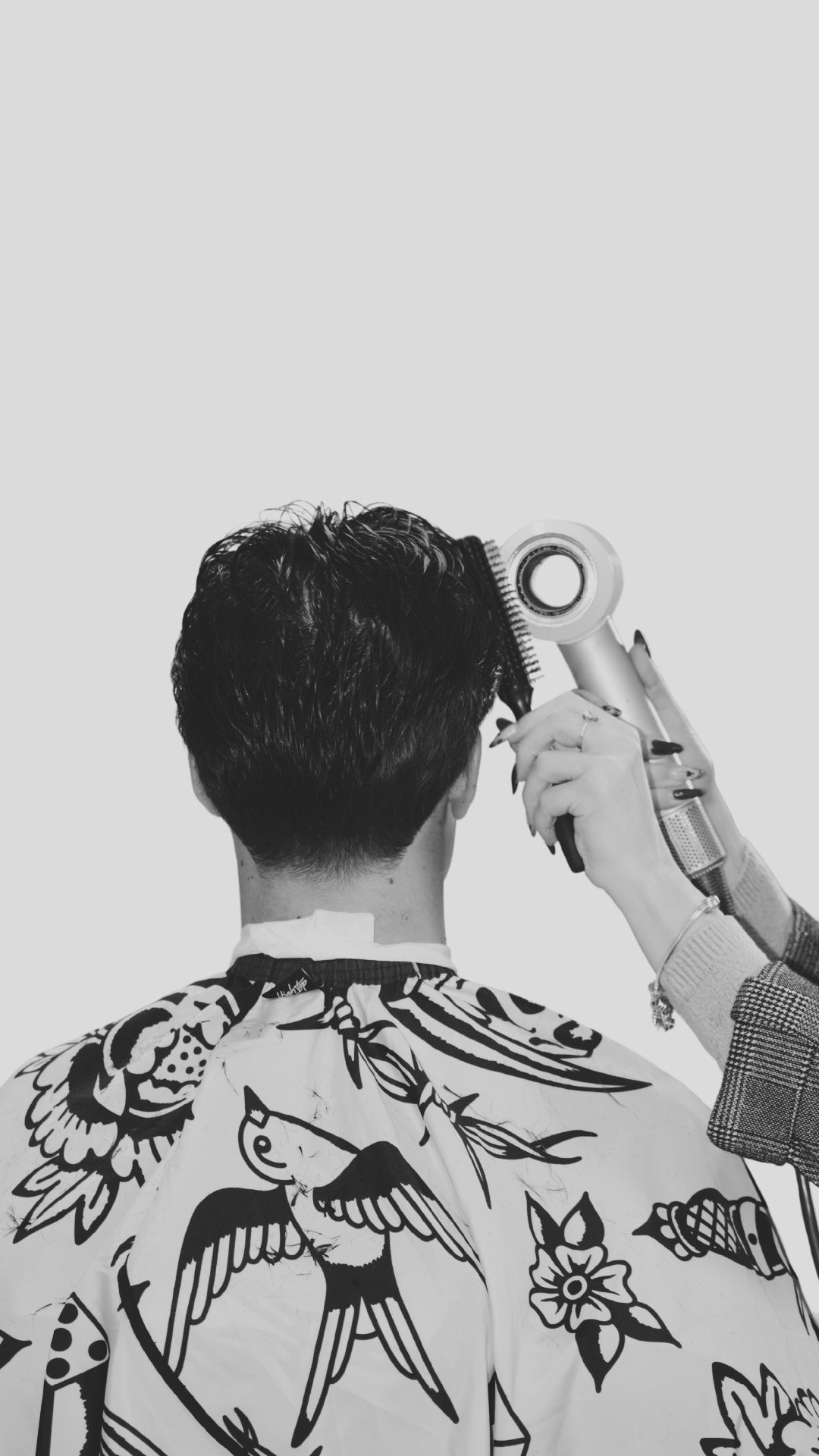 A woman styling a man's hair with a hairdryer in a salon, seen from the back, wearing a cape with a tattoo-style eagle and floral designs.