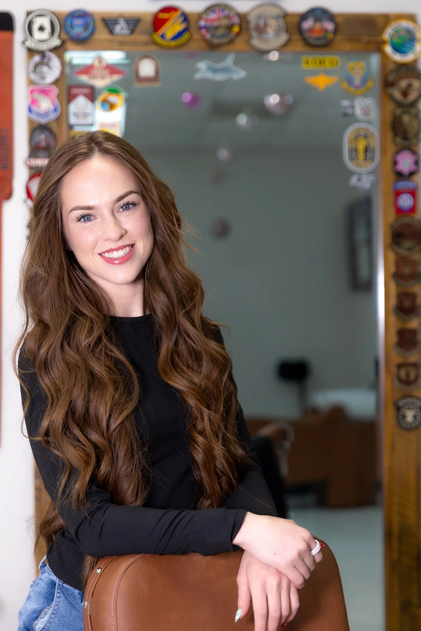 A young woman with long, wavy auburn hair and blue eyes sitting on a brown chair, smiling at the camera. She is wearing a black long-sleeve top and light blue jeans. In the background, there is a framed mirror decorated with various patches and badges.