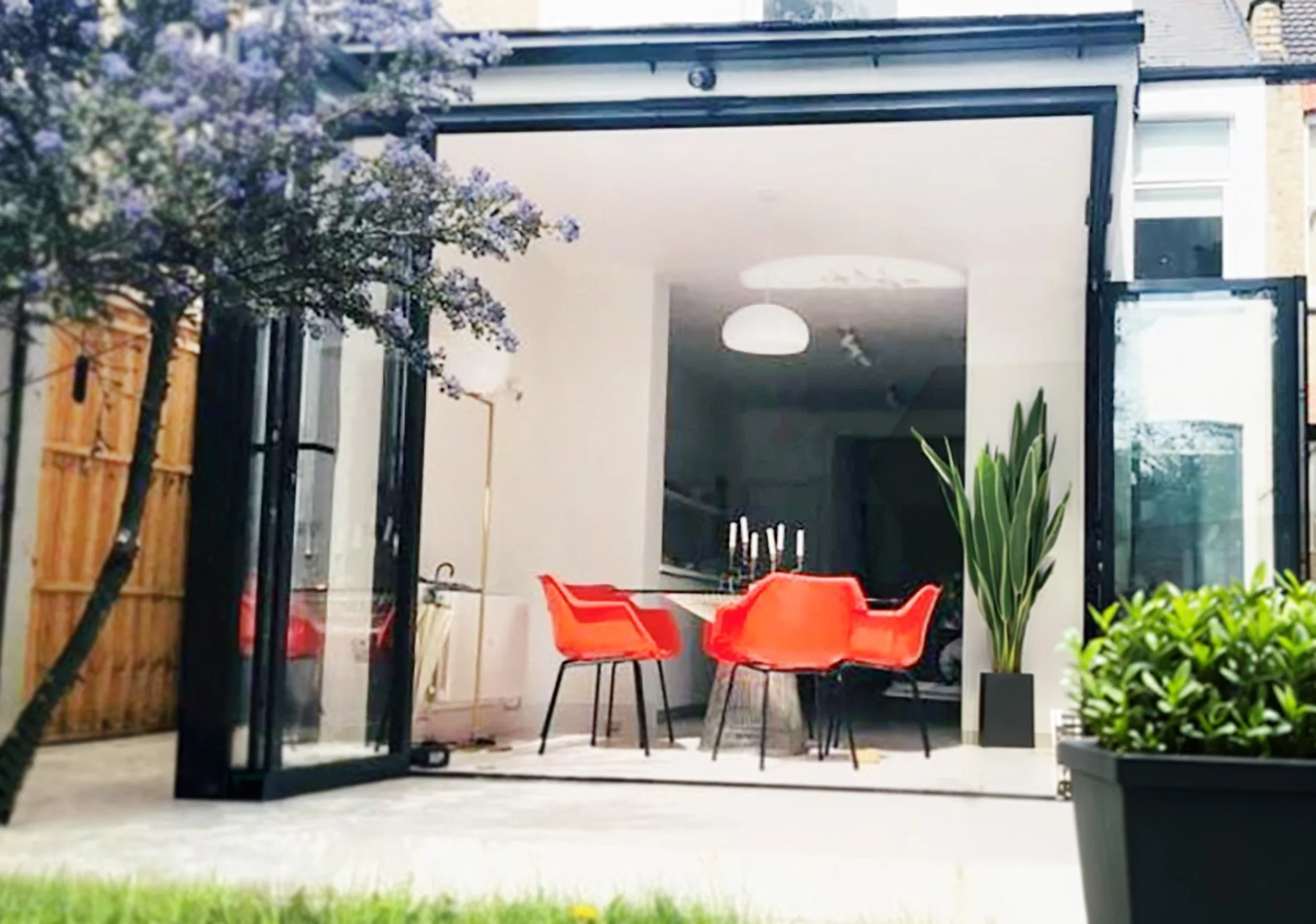 A modern outdoor patio with a dining area featuring a round table and four orange chairs, a glass door opening into the house, green plants, purple flowering bonsai, and a wooden privacy fence.