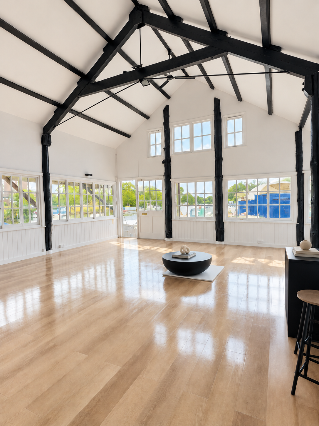 Spacious open-plan interior refurbishment featuring vaulted ceilings, exposed black timber beams, large windows, white panelled walls and light oak flooring.