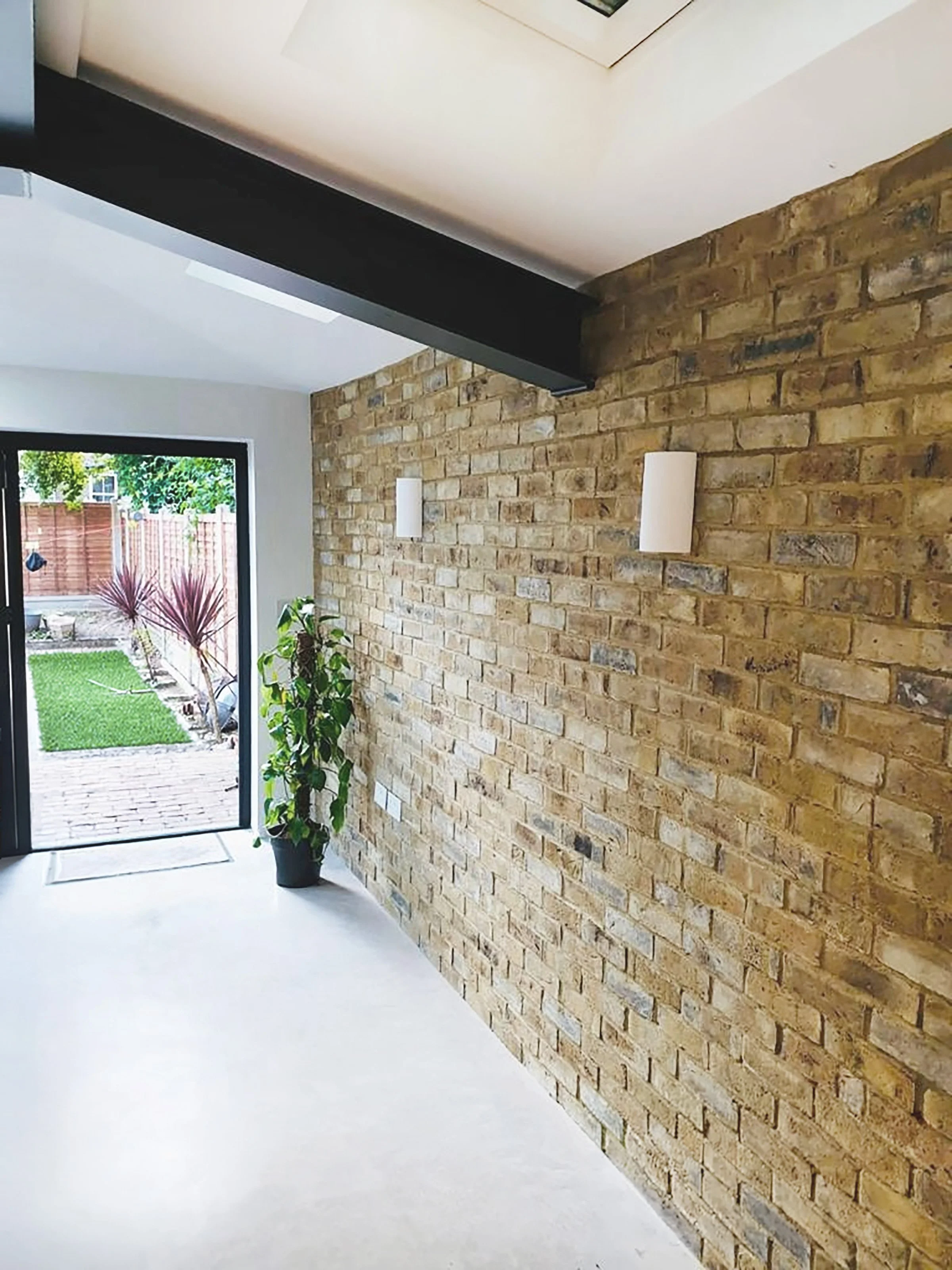 Interior view of a bright room with a brick wall, white ceiling, and large glass door leading to a garden with a green lawn, potted plants, and outdoor trees.