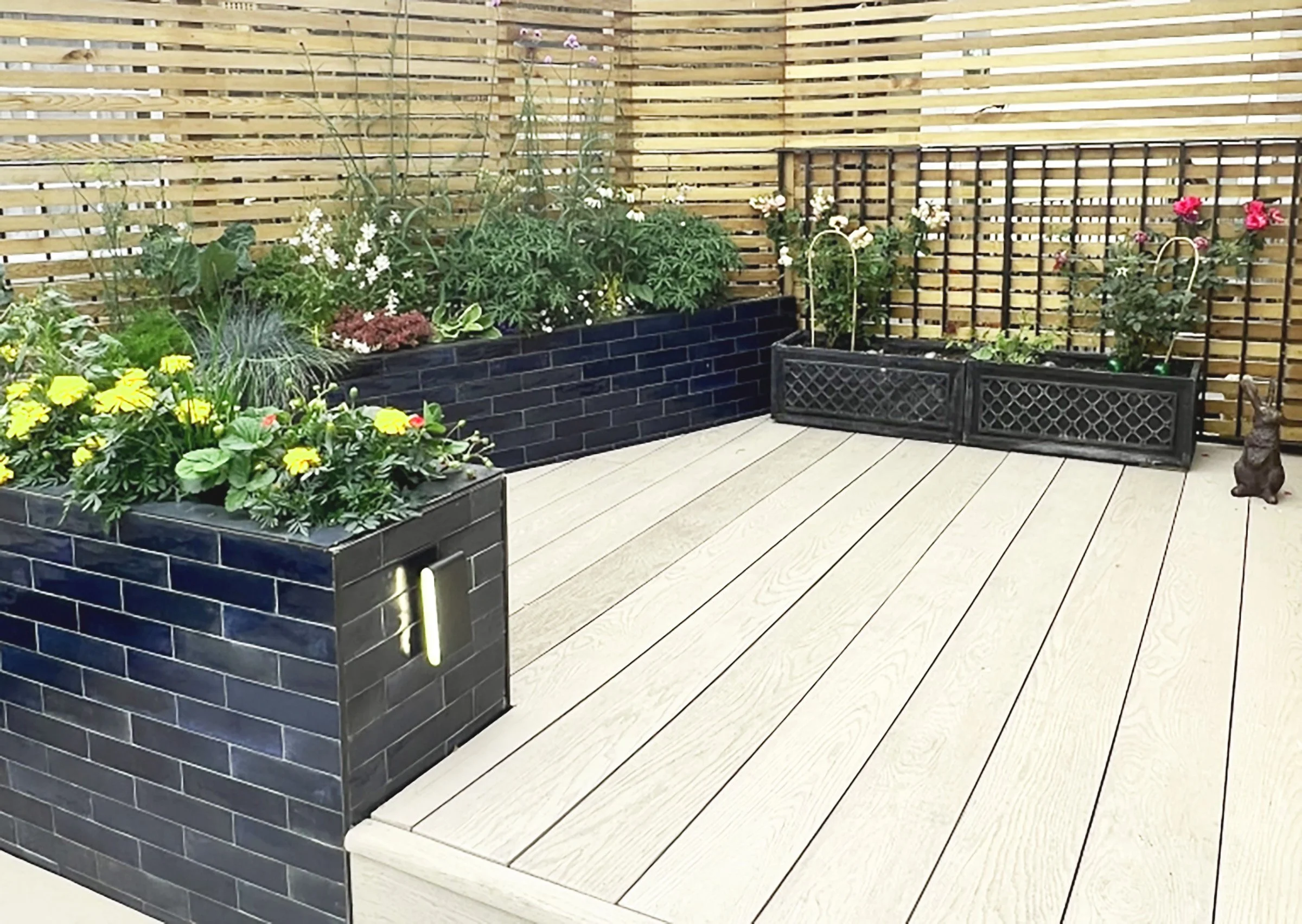 Decorative outdoor space featuring wooden decking, black brick planters with colorful flowers, grid fencing, and a small animal figurine.