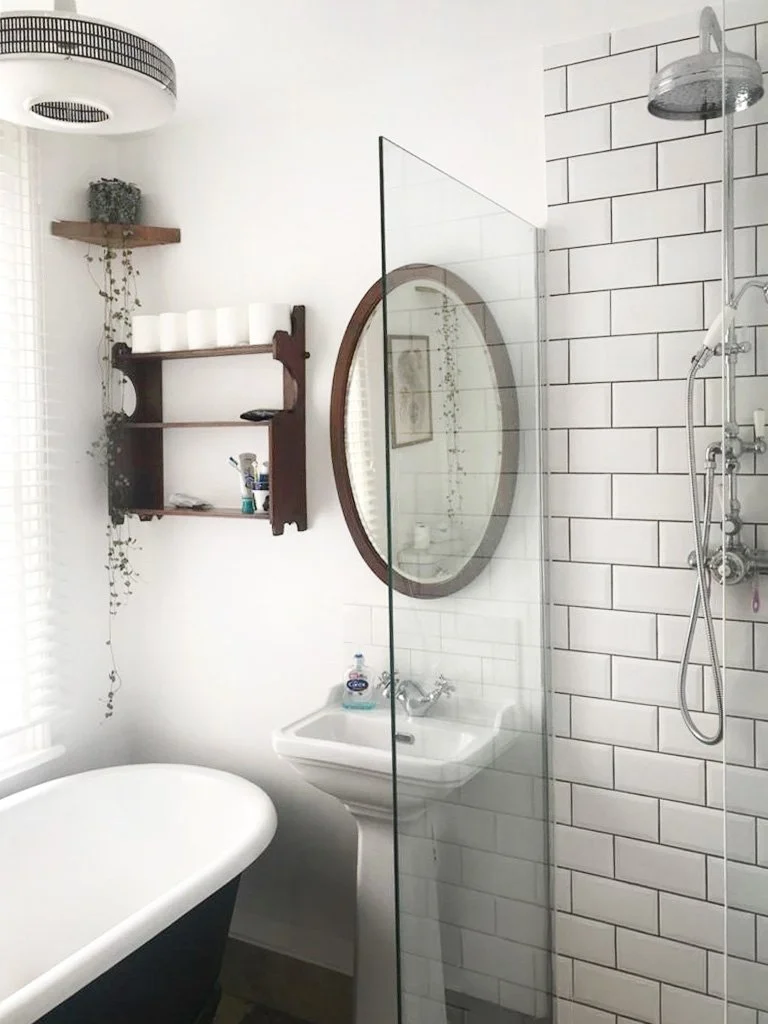 A bathroom with a clawfoot bathtub, a small pedestal sink, and a walk-in shower with white subway tiles. There is an oval mirror above the sink, a wooden shelf with toiletries, and a modern ceiling light fixture.