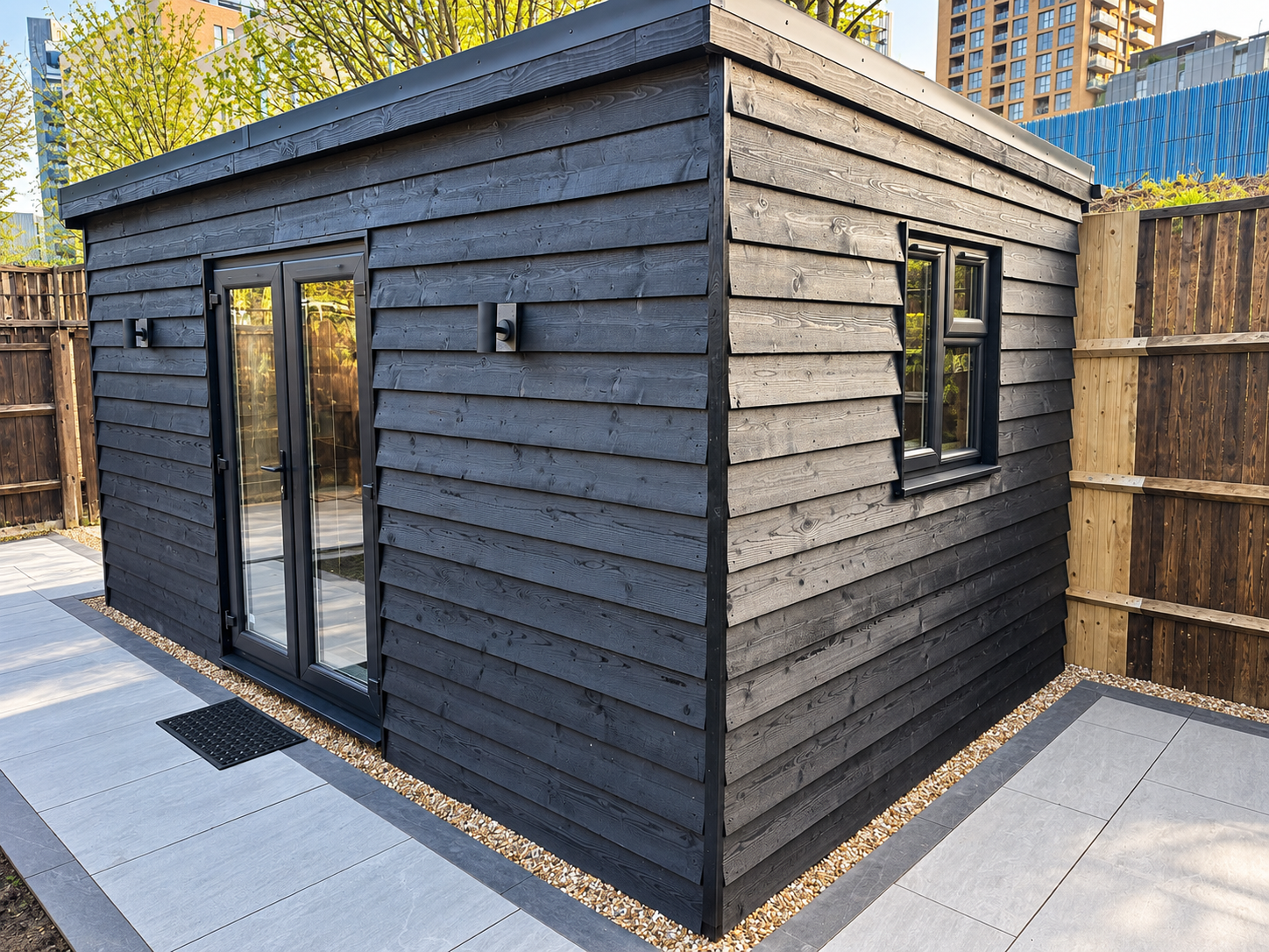 Modern bespoke garden room in Guildford featuring dark timber cladding, black-framed French doors, double-glazed windows and contemporary grey patio paving.