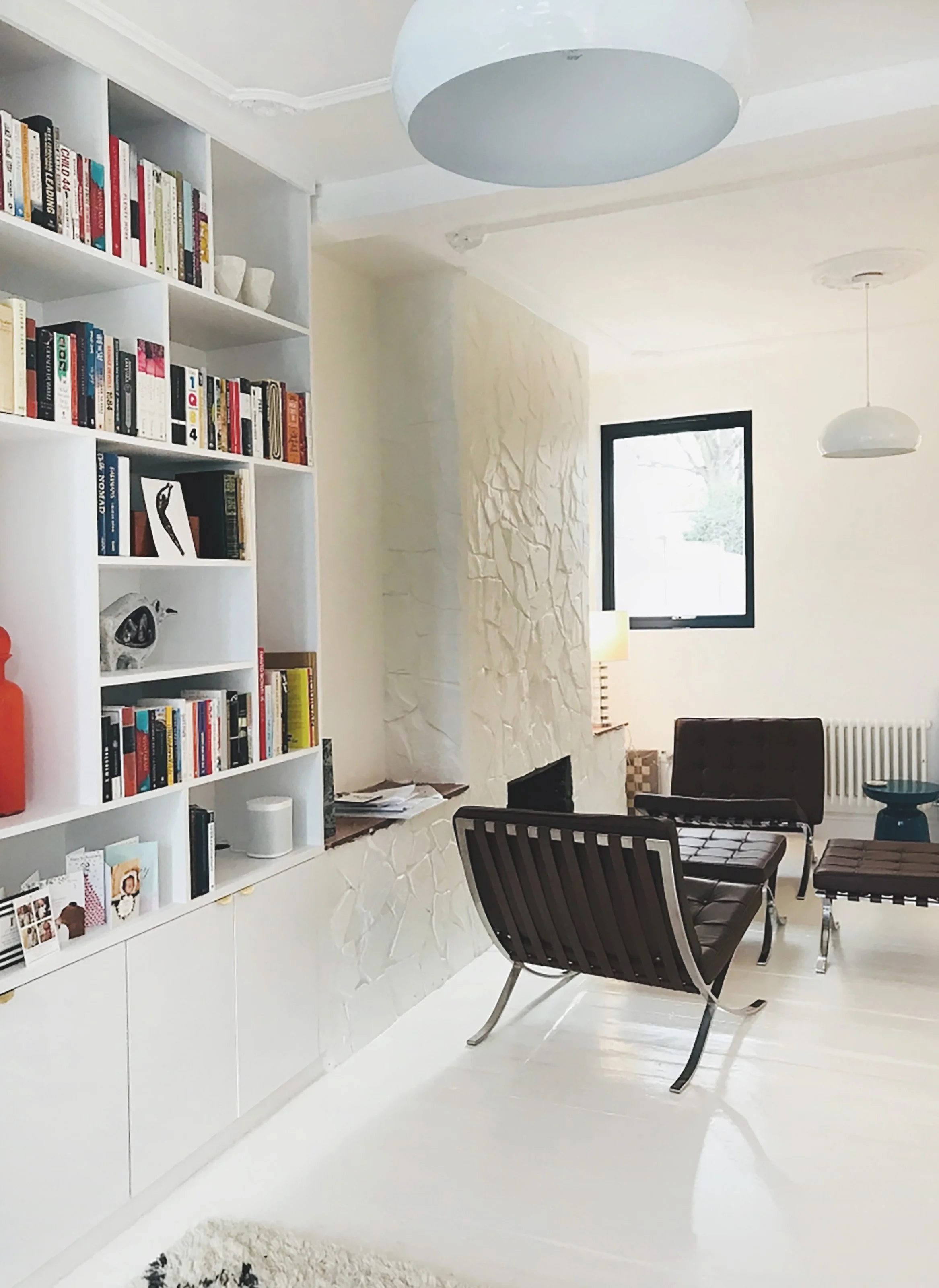 Modern living room with white bookshelves filled with books and decor, white textured wall, black-framed windows, black chairs with metal legs, and a small dark blue table. Bright and minimalist design.
