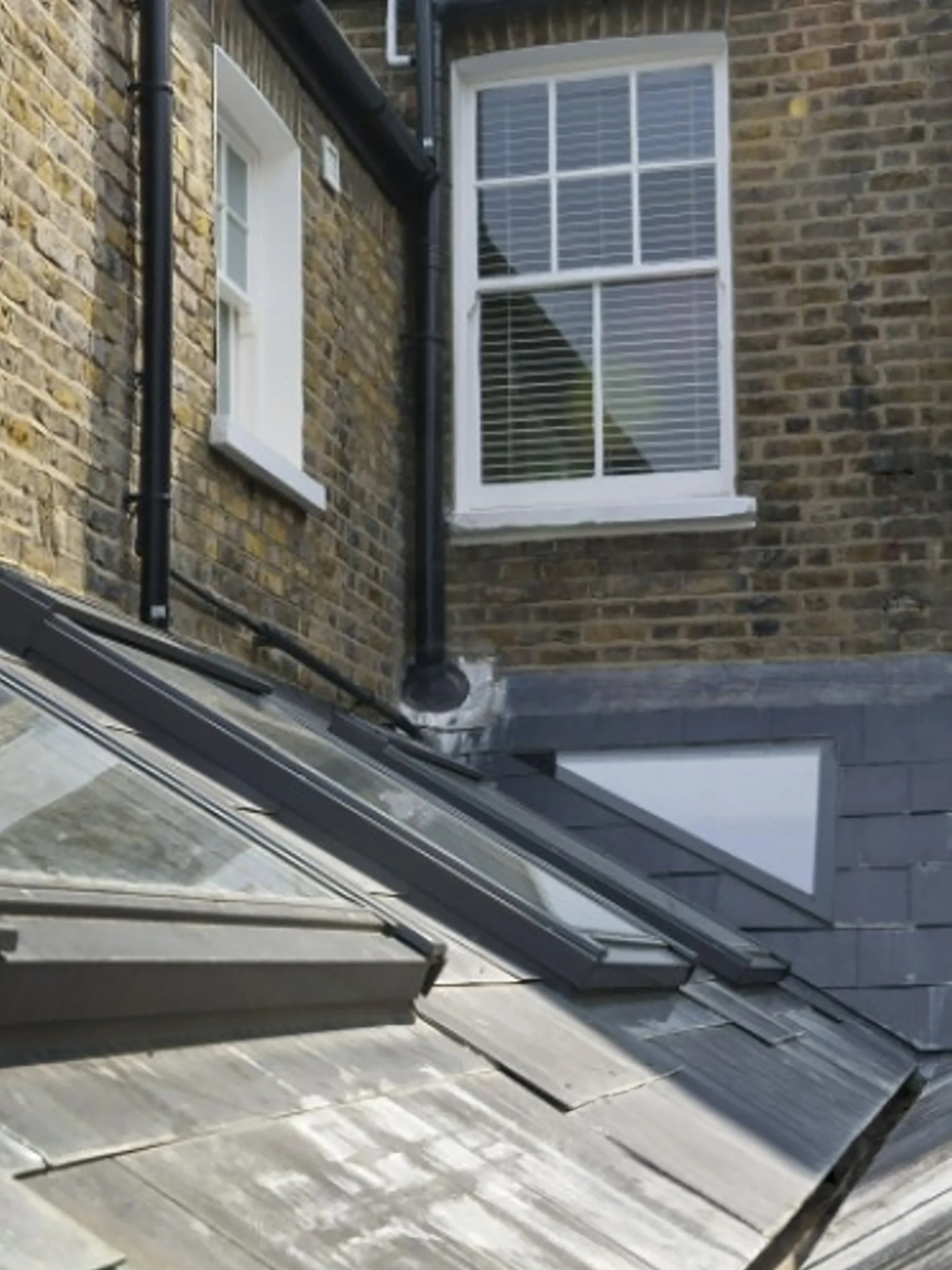 View of a brick building with white-framed windows, black drainpipes, and a glass skylight on the roof seen from inside a conservatory or patio area with glass panels.
