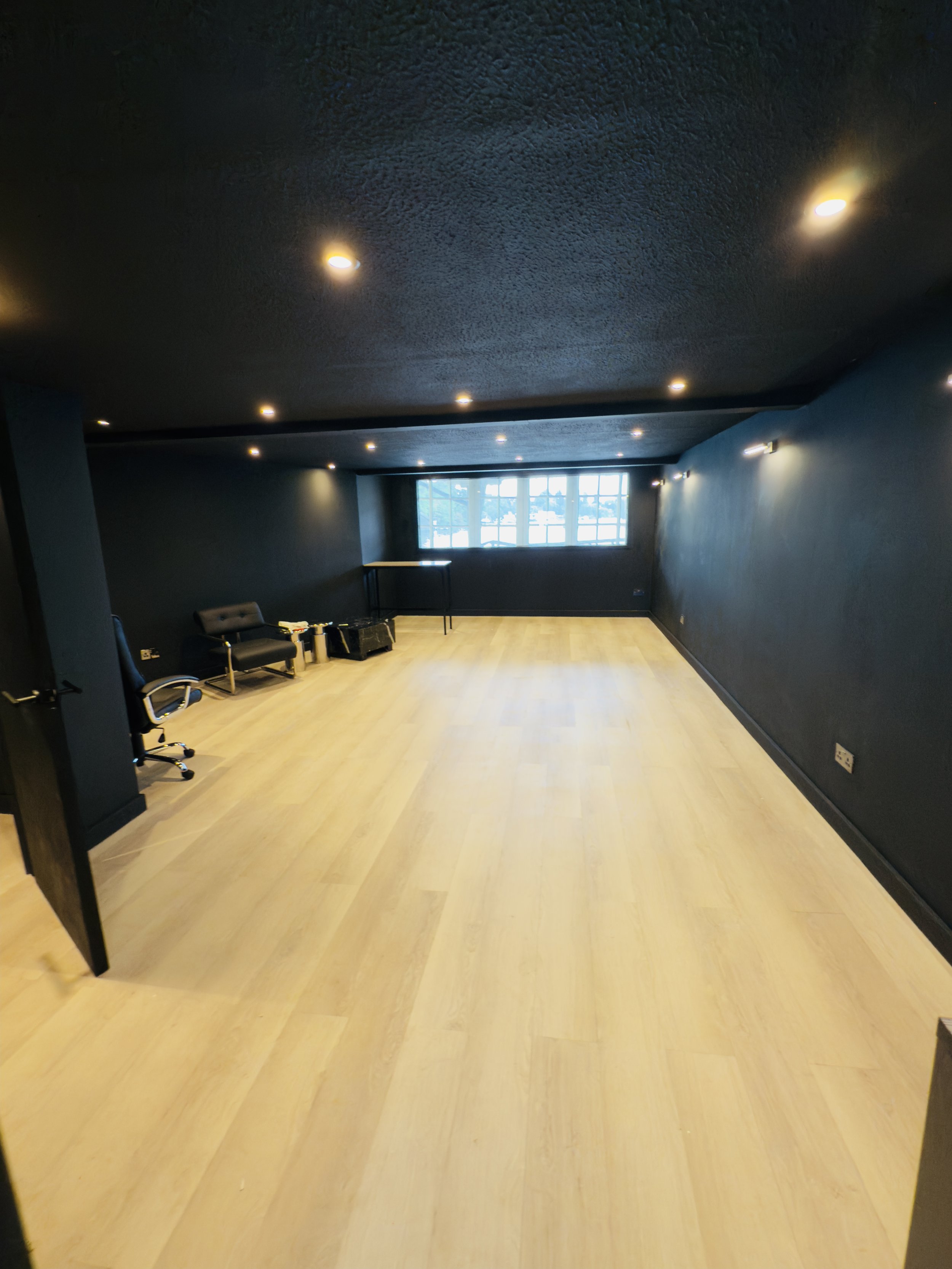 A bold, contemporary renovation pairing matte black ceiling and walls with warm light oak laminate flooring for striking contrast. Recessed spotlights and wall-mounted strip lighting layer the space with light, while a large crittall-style window bri