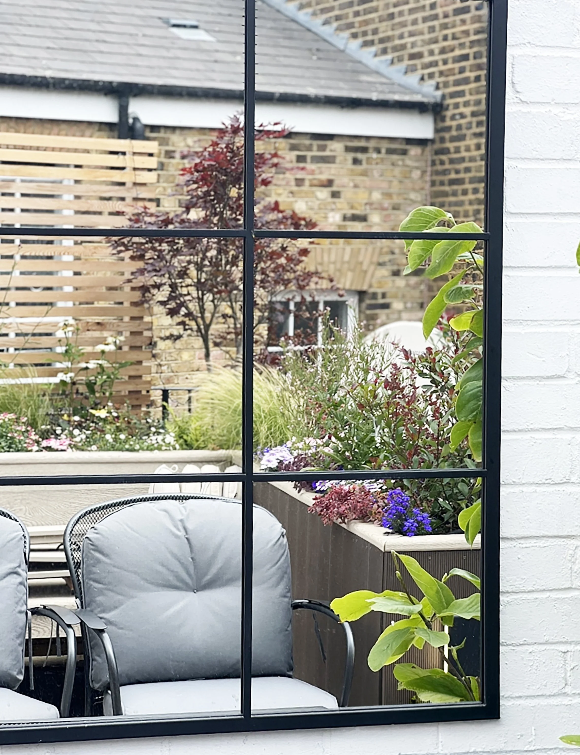 View through a window showing an outdoor patio with gray chairs, a flower bed with purple, pink, and white flowers, a wooden fence, a tree with reddish leaves, surrounding houses with brick and stone walls, and some green plants.