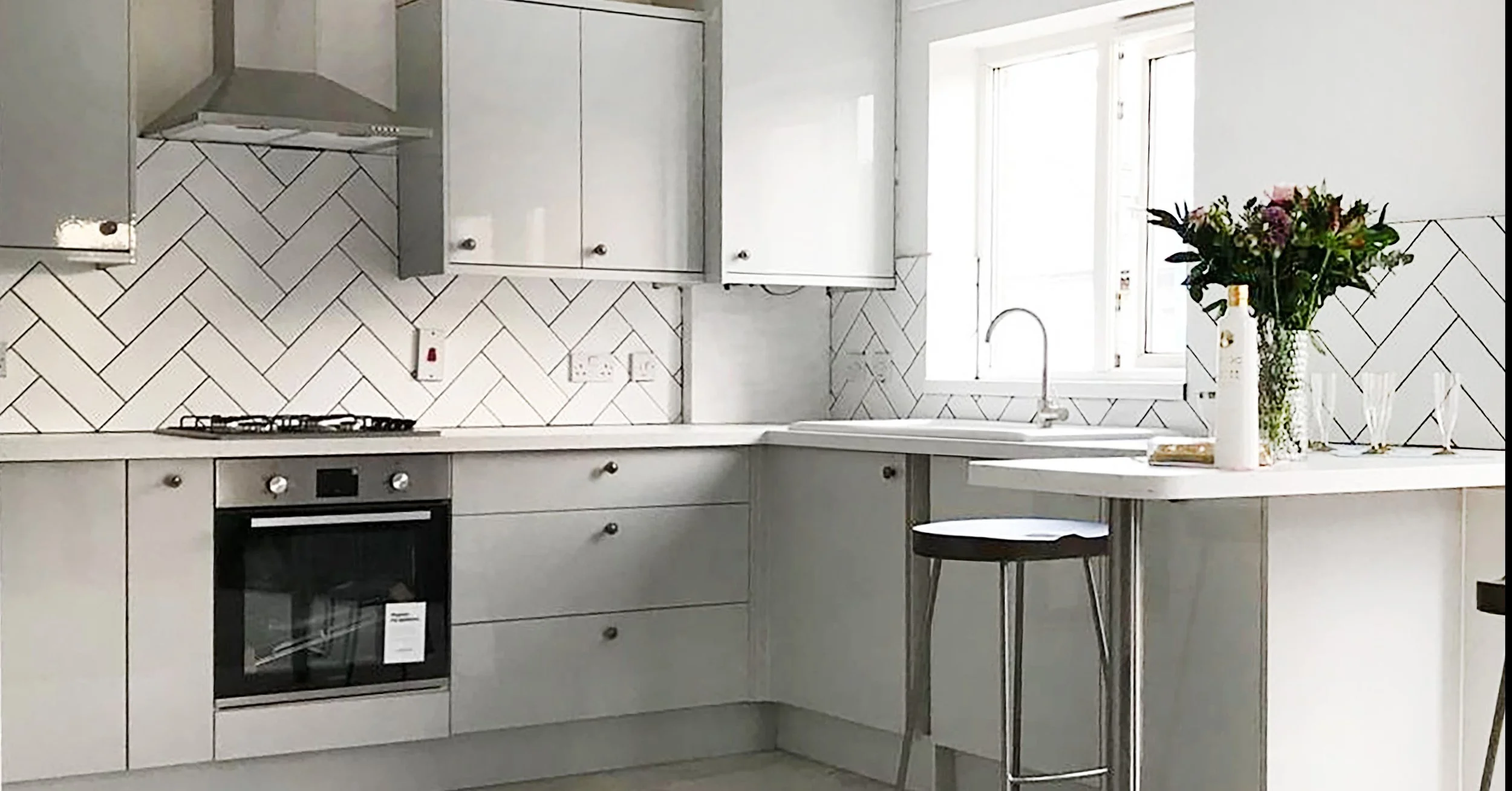 A modern kitchen with white cabinets, a gray stove, white countertop, a window with natural light, a vase of flowers on the counter, and a small round black stool.