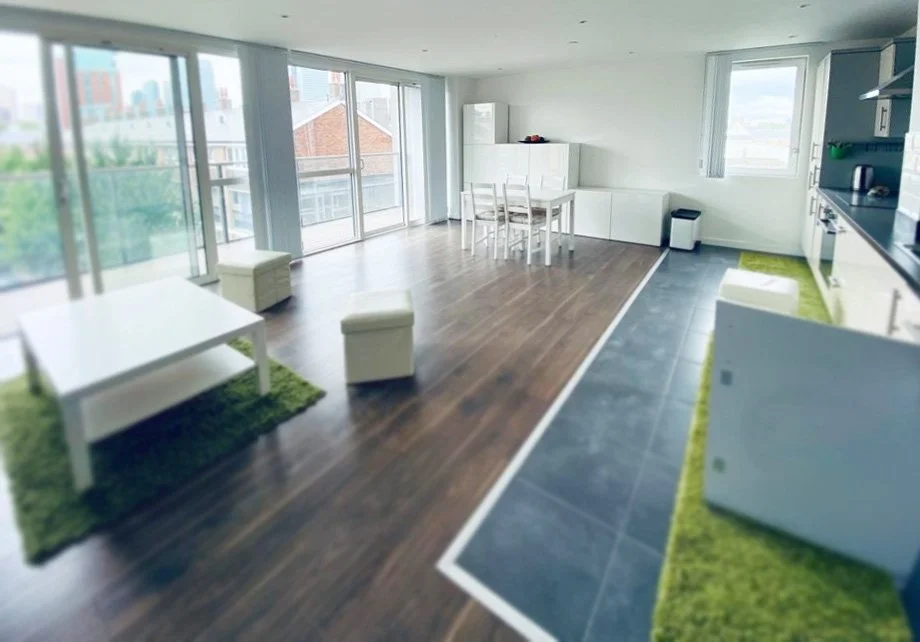 Spacious apartment living room with large windows, a white coffee table on a green rug, two white ottomans, a small dining table with chairs, and a kitchen area with dark tiles and white cabinets.