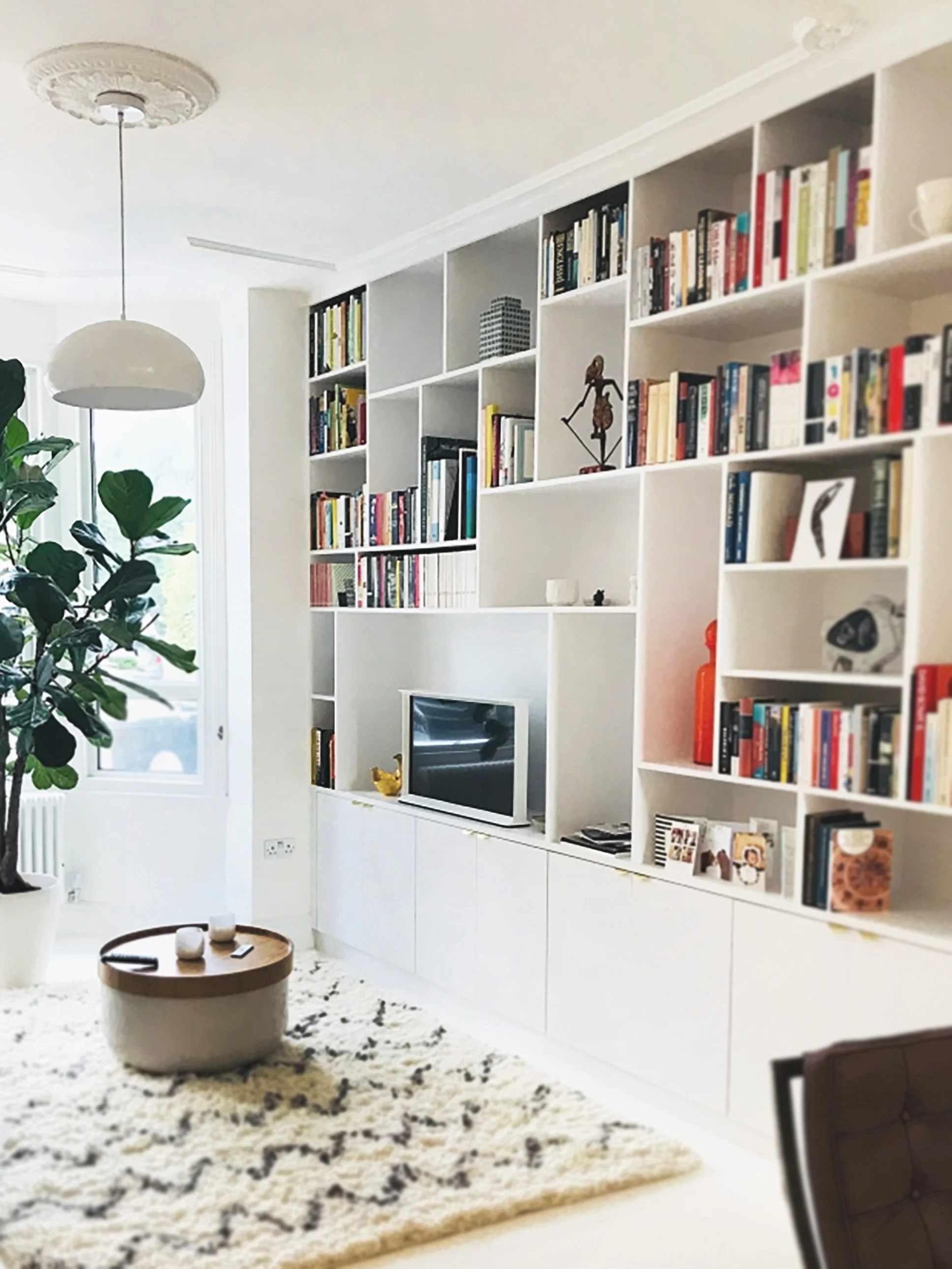 Living room with white shelving unit filled with books, a TV, decorative items, a green plant, and a round coffee table on a cream and black patterned rug.