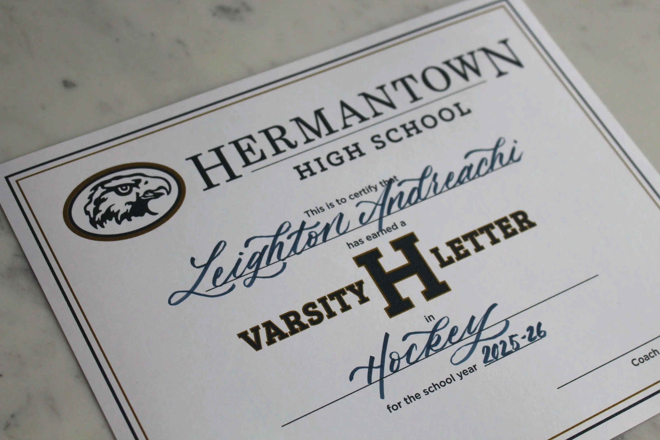 A school varsity letter sits on a marble surface with a calligraphy script that says "Leighton Andreachi" and they lettered in Hermantown Hockey.