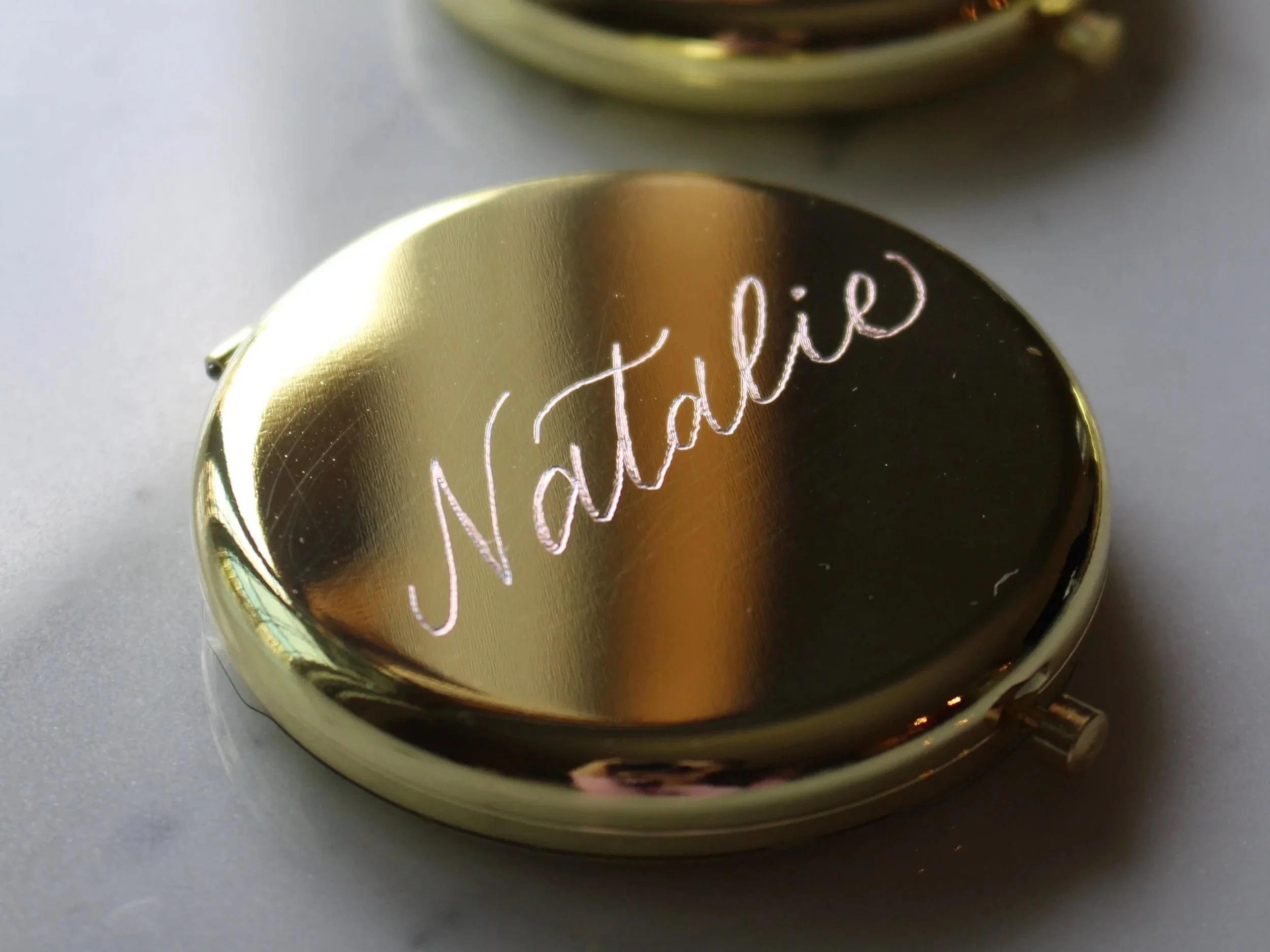 Close-up of a shiny, gold-colored mirror compact with the inscription 'Natalie' in cursive on its surface, lying on a marble background.