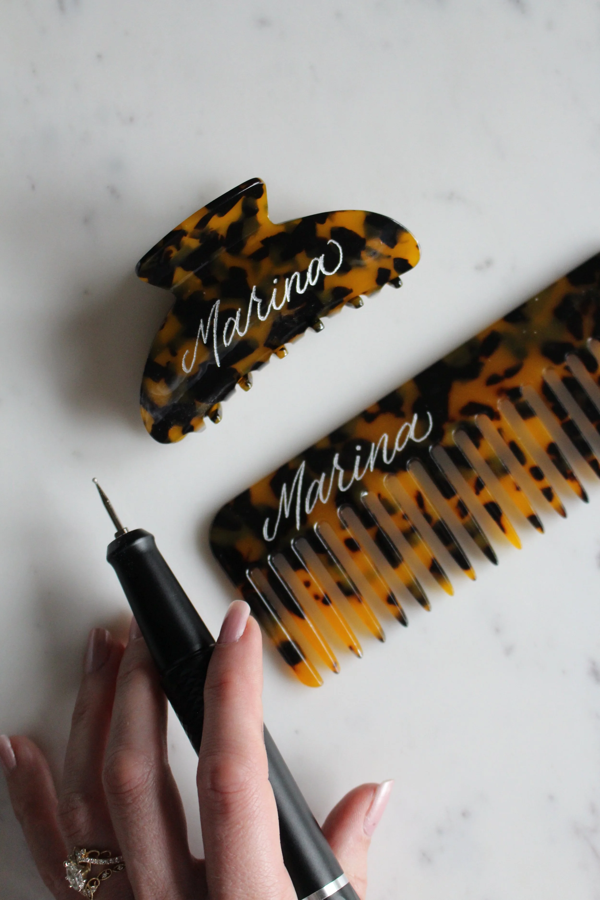 Custom engraved  tortoise shell comb and hair claw clip sit on a marble table with the name “Marina” inscribed on both items.