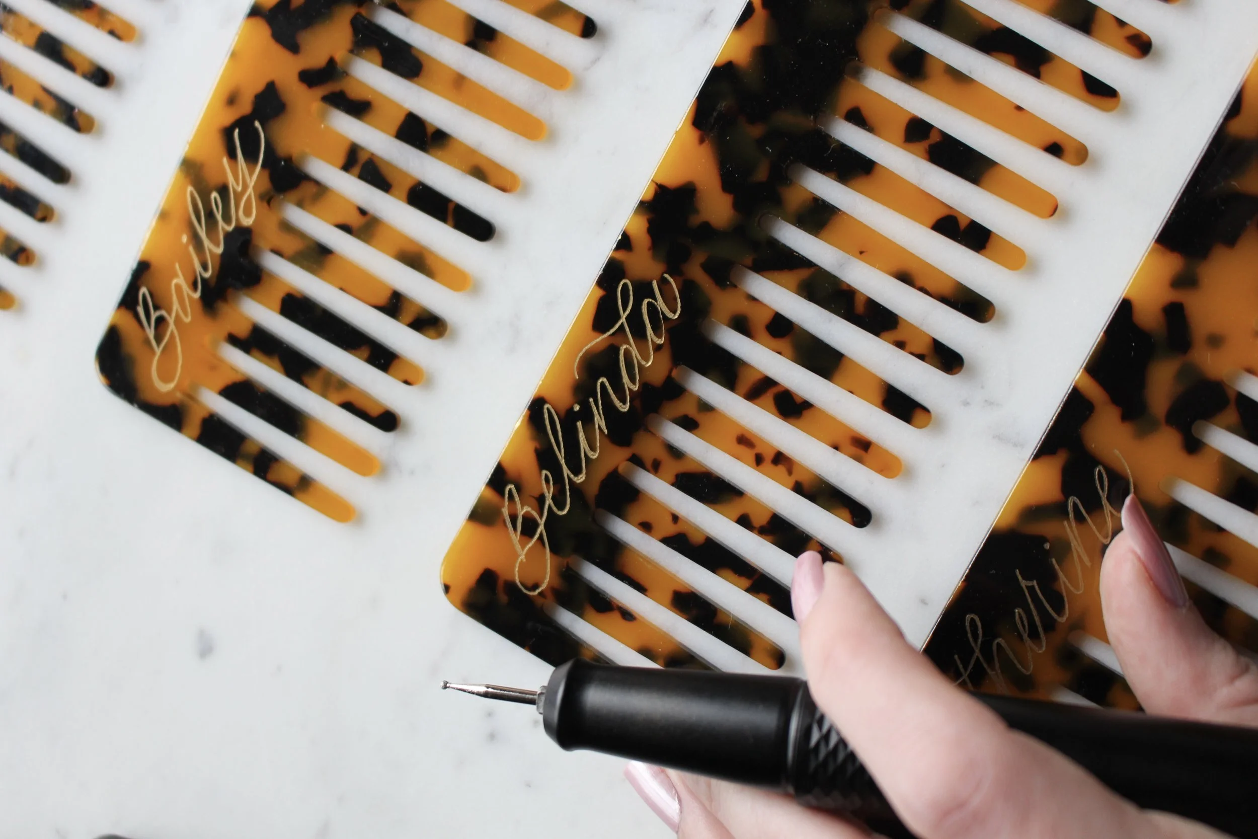 Hand Engraved Tortoise Shell Combs with beautiful calligraphy on them that say "Bailey" on one comb and "Belinda" on the other. Belle has her hand slightly in the frame displaying her tool she uses to engrave the combs.