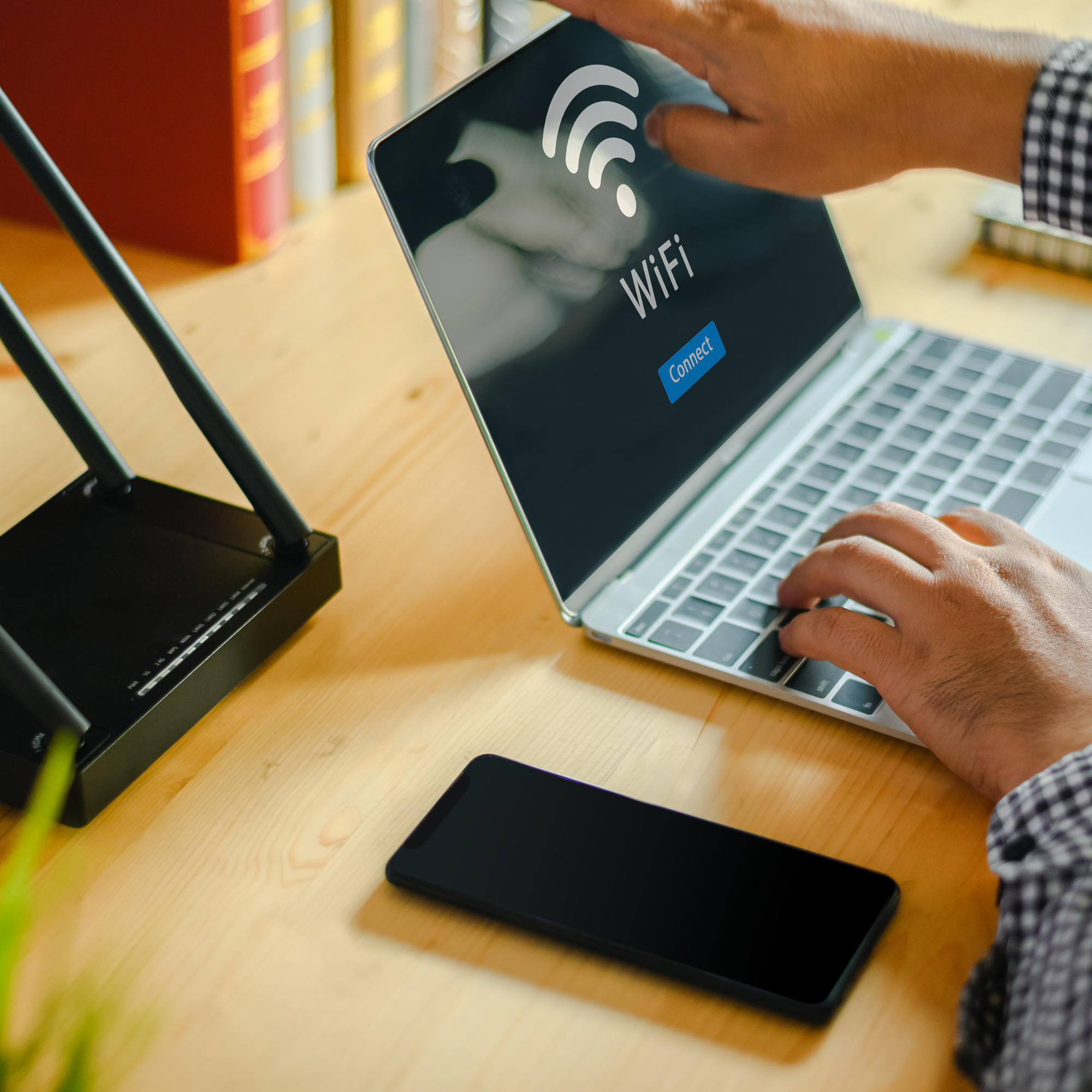 A person's hand hovering over a smartphone with a Wi-Fi connection screen visible. The phone is placed on a wooden desk with books in the background and a silver laptop partially visible.