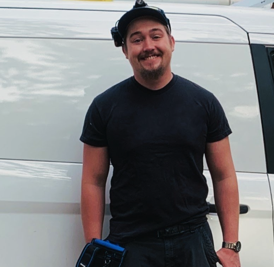 Man smiling and wearing a black shirt, standing outdoors next to a white van, holding a small device in his right hand, wearing a watch and a headlamp.