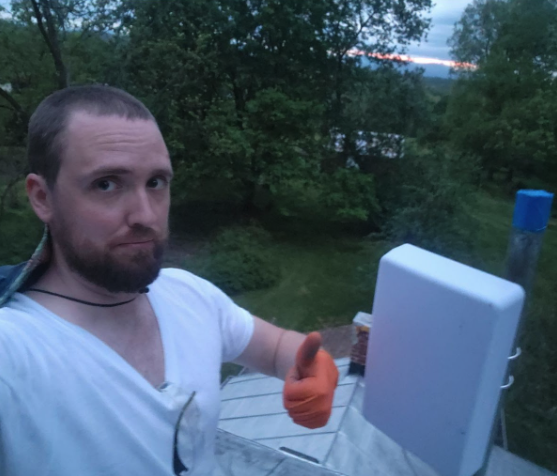 Man with short hair, beard, and white t-shirt giving a thumbs up in an outdoor setting with trees and greenery in the background, near some equipment or appliances.